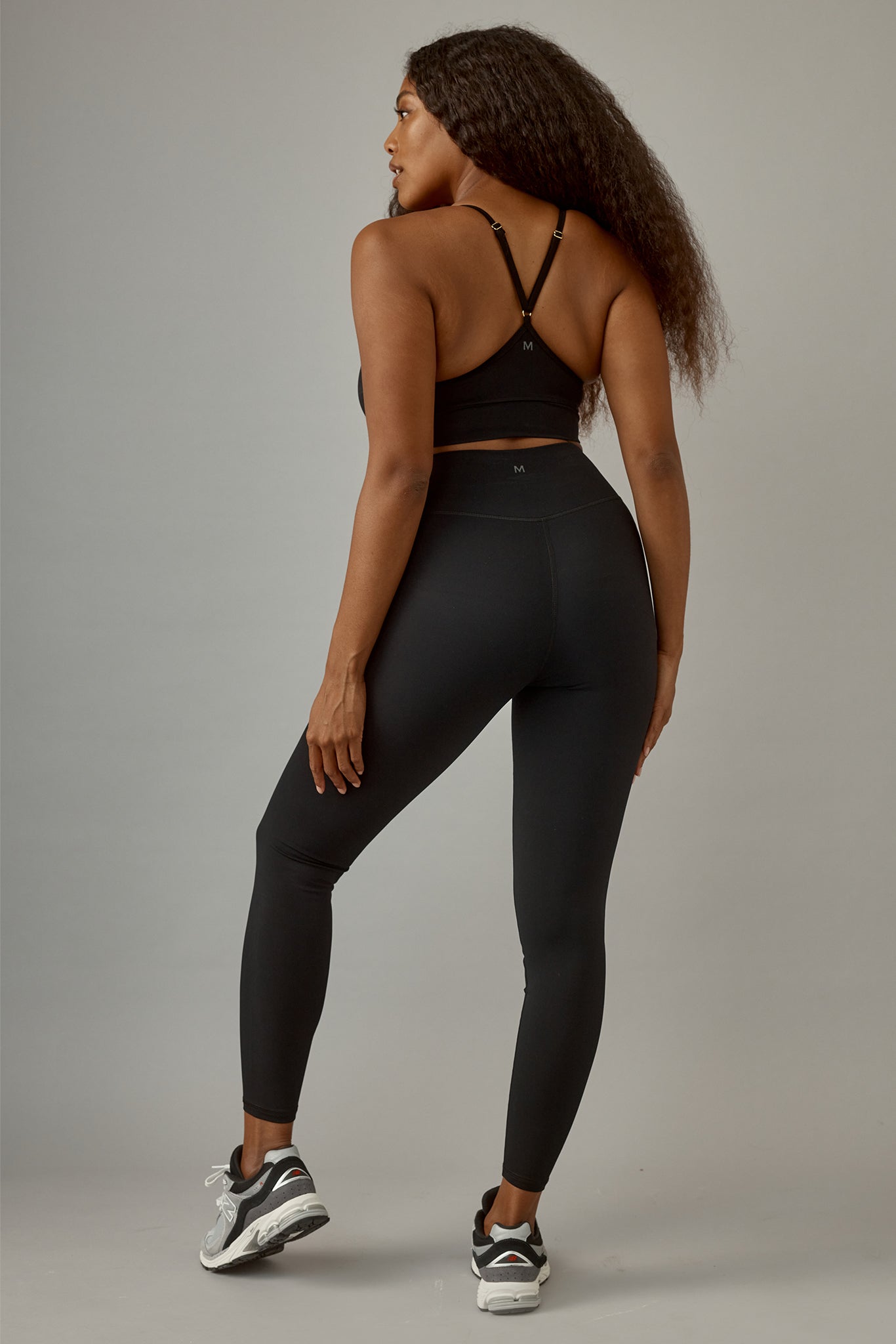 Woman facing away in black racerback sports bra and high-waist leggings, small M logo on bra and waistband, wearing sneakers.