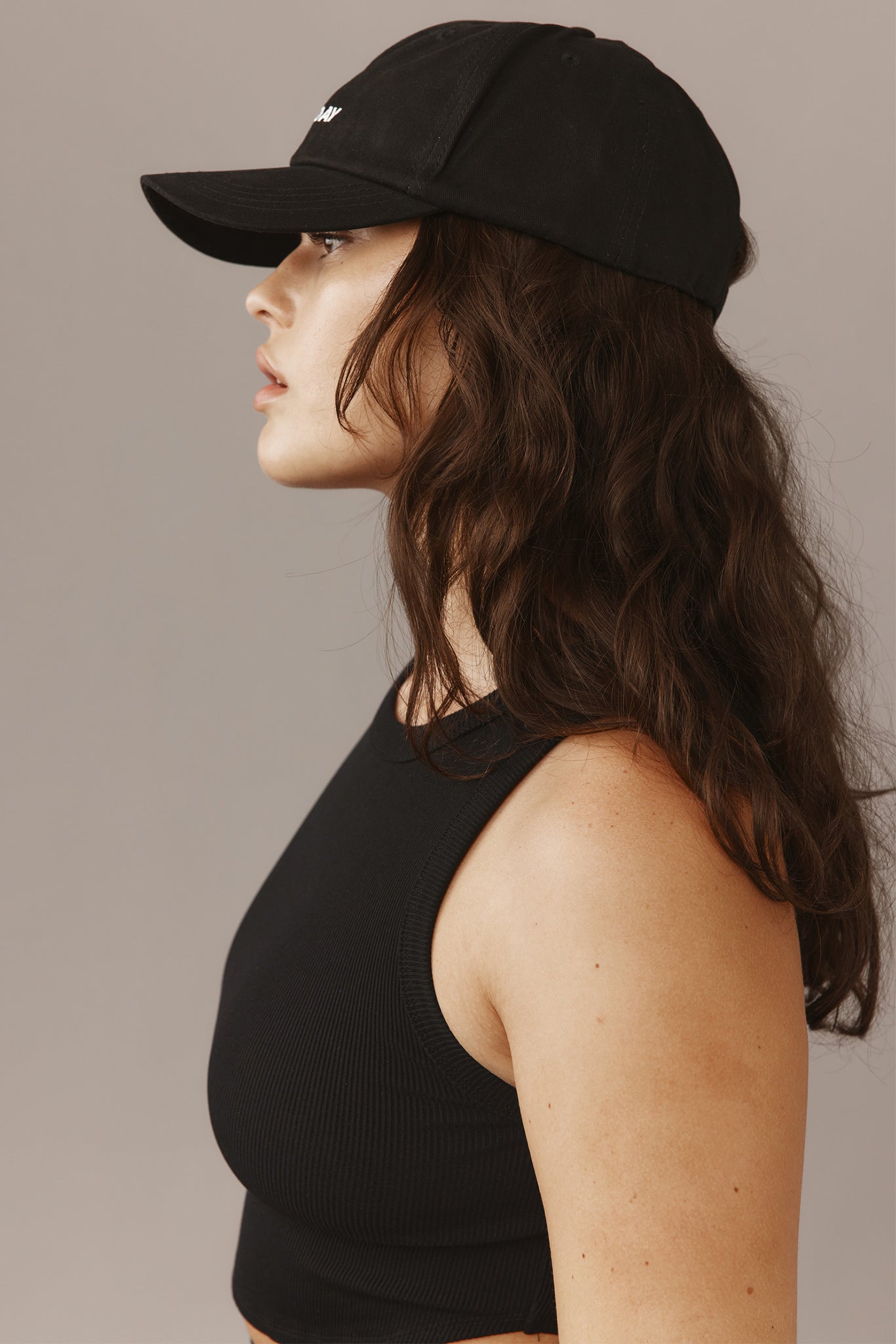 Woman in profile wearing black baseball cap embroidered "DAY", black ribbed sleeveless top, long wavy brown hair, neutral background.