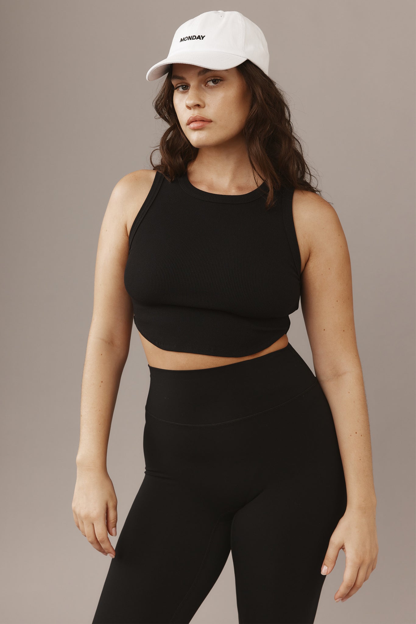 Woman wearing a white baseball cap labeled MONDAY, black cropped tank and high-waist leggings, neutral background.