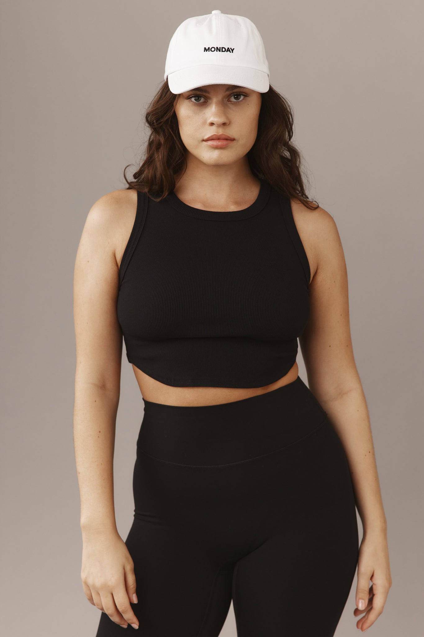 Woman facing camera wearing a black cropped tank top, high-waist leggings, and a white cap embroidered 'MONDAY'.