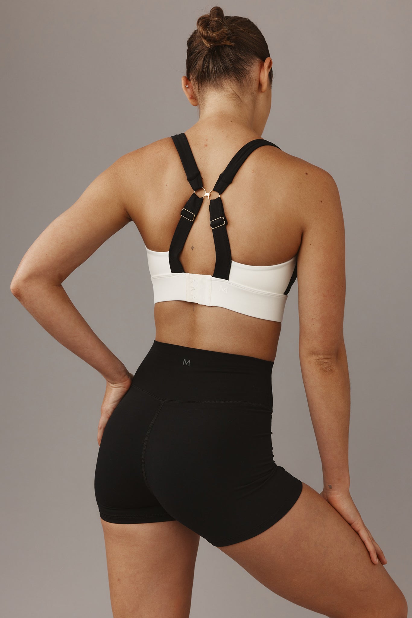 Rear view of a woman wearing a white sports bra with crossed black straps and black high‑waist shorts labeled M.