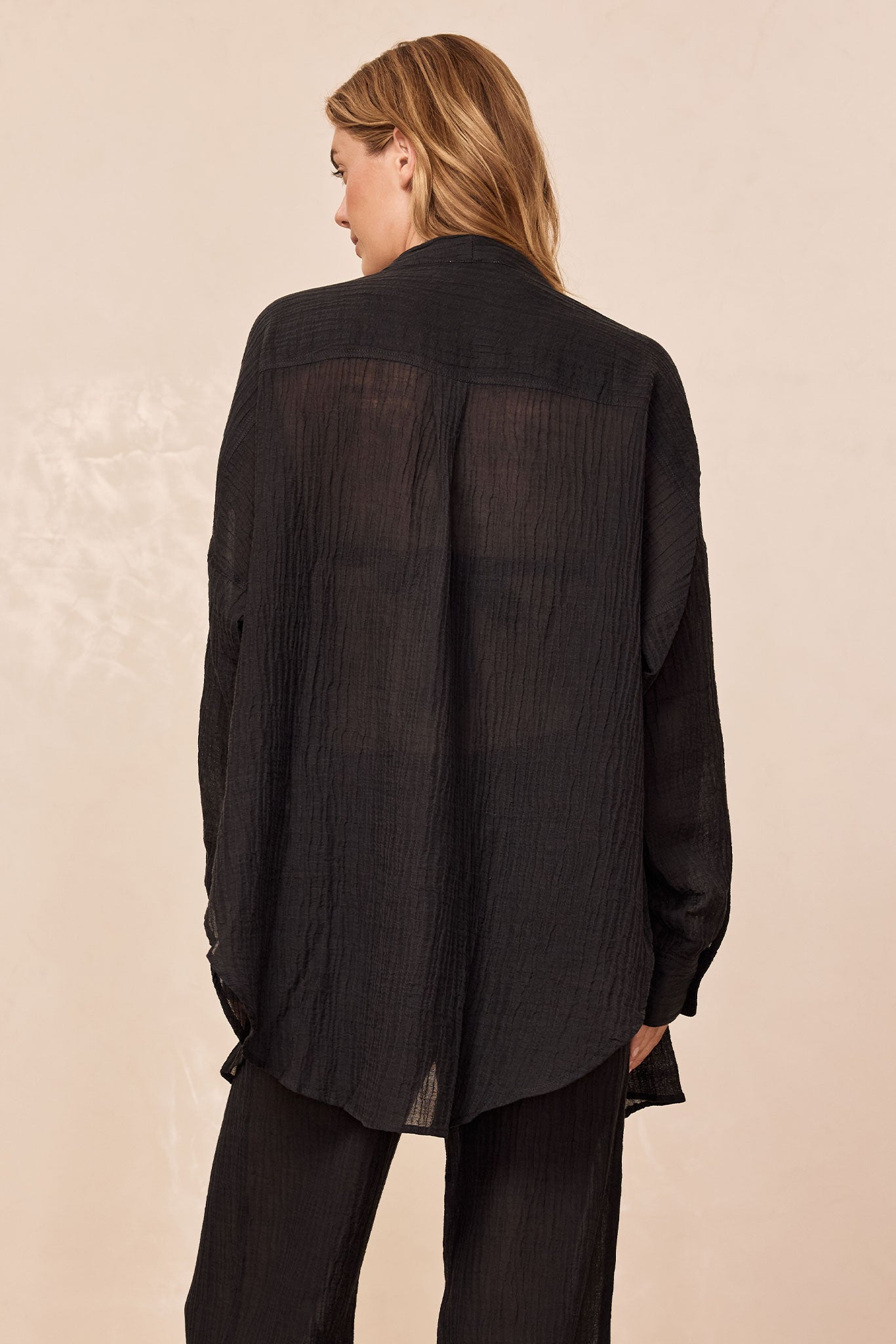 Model shown from back wearing a sheer, textured black long-sleeve shirt with matching black wide-leg pants.