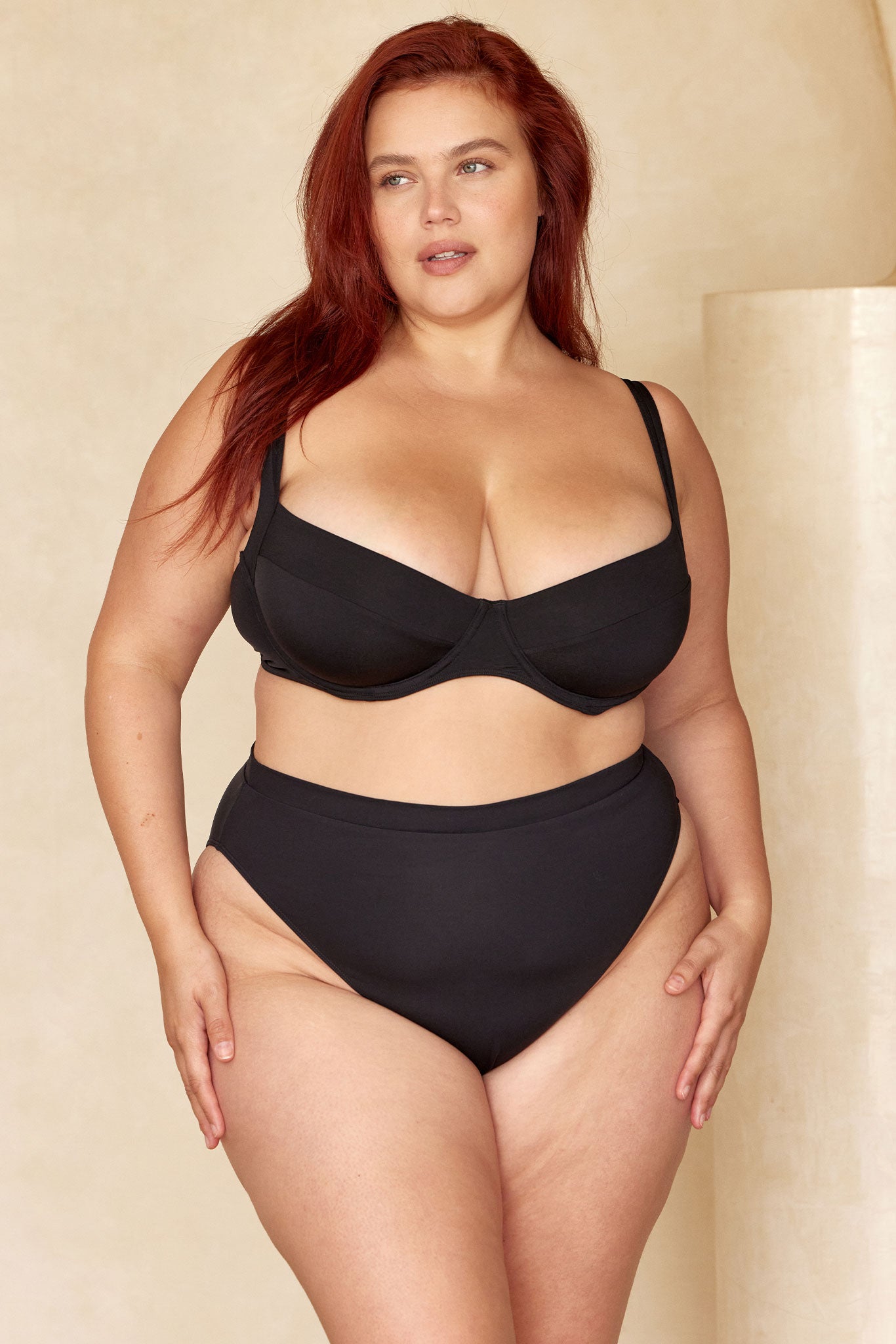 Plus-size model with red hair in a black underwire bikini top and high‑waist bottoms, standing against a beige backdrop.