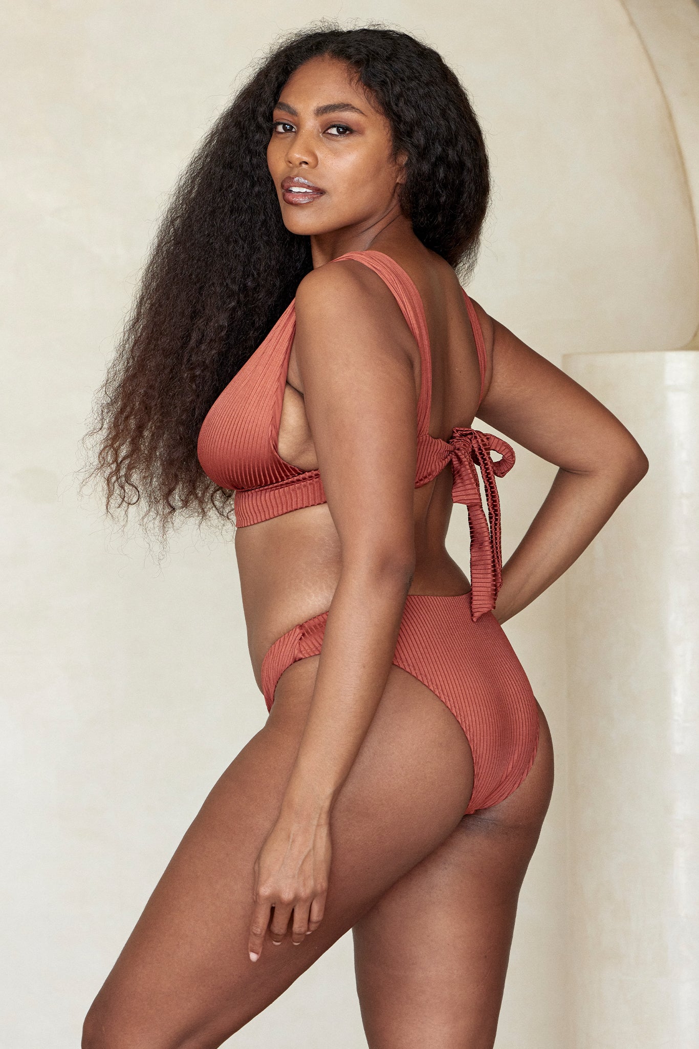 Woman with long curly hair wearing a rust ribbed bikini, three-quarter back pose, looking over shoulder, hand on hip.
