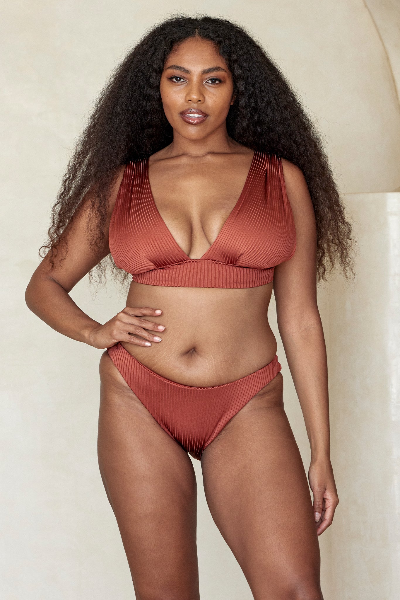 Woman model wearing rust-colored ribbed bikini, standing front view with hand on hip against a neutral background.