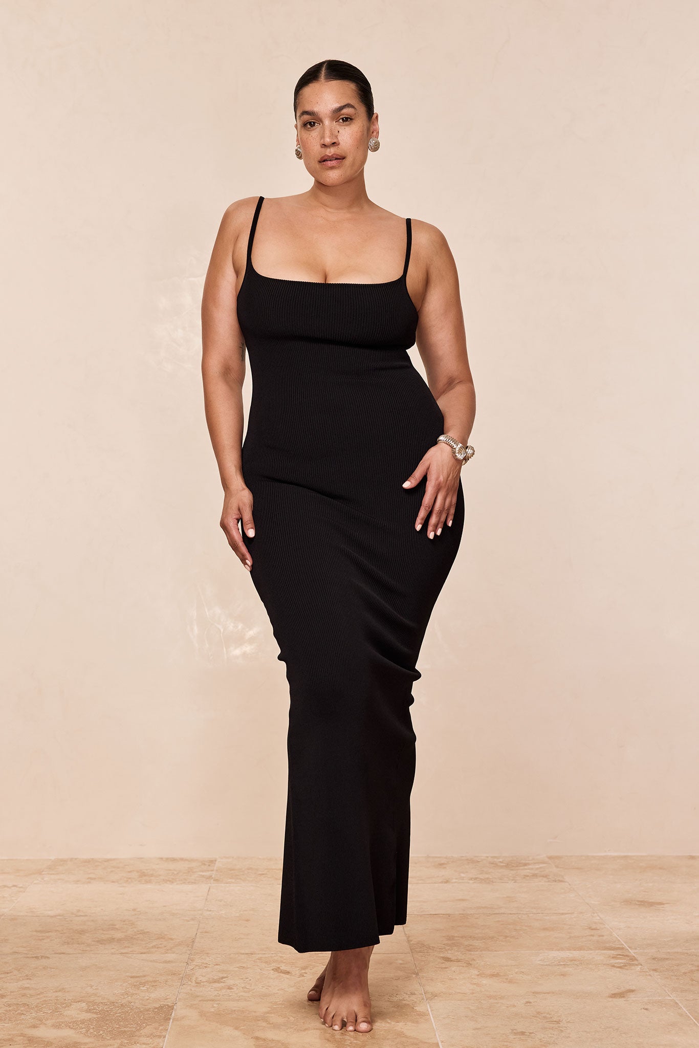 Woman wearing fitted black ribbed maxi dress with thin straps, bare feet, earrings and bracelets, standing on beige tile.