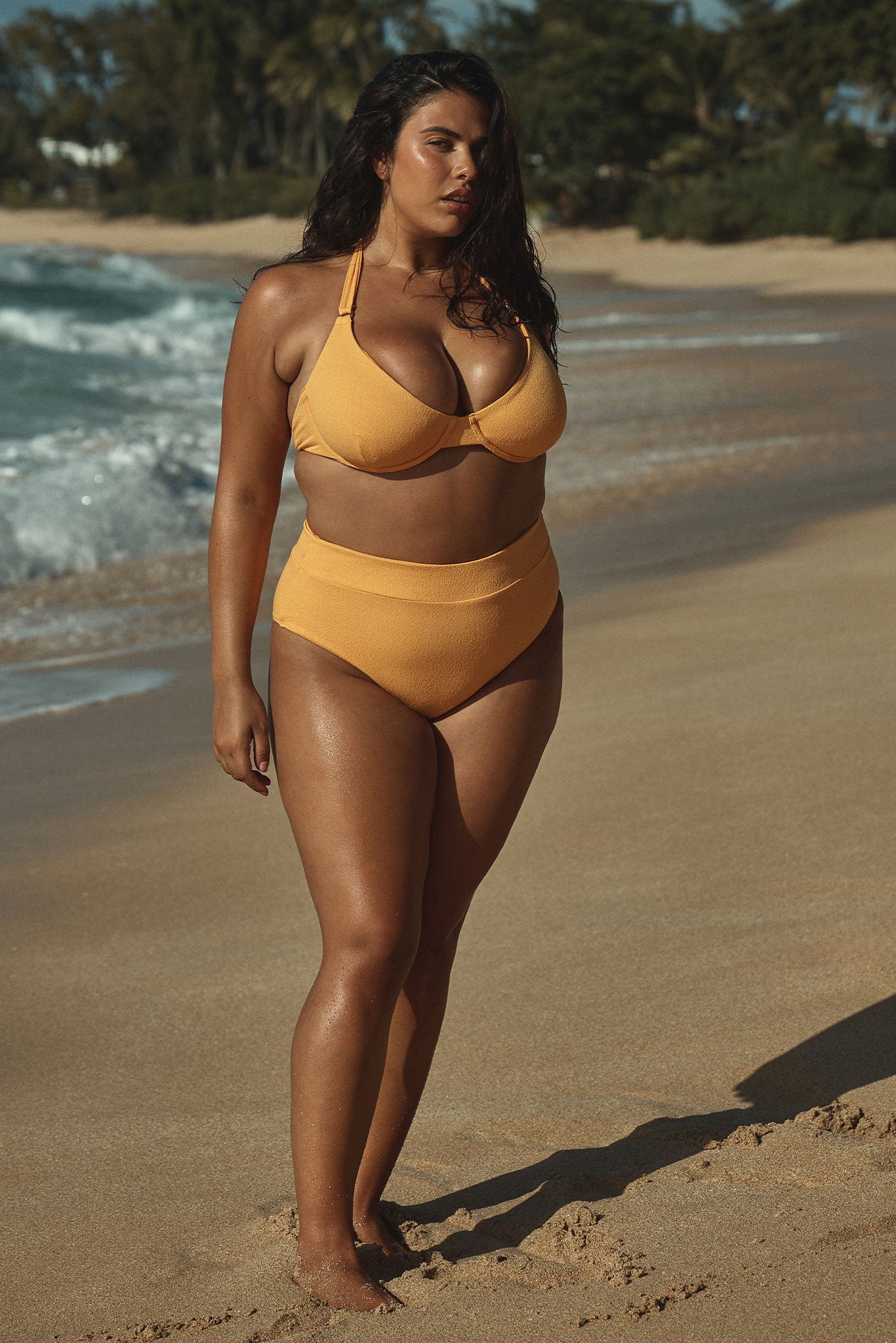 Plus-size model wearing a yellow bikini standing on a sandy beach near waves, facing slightly to her left.