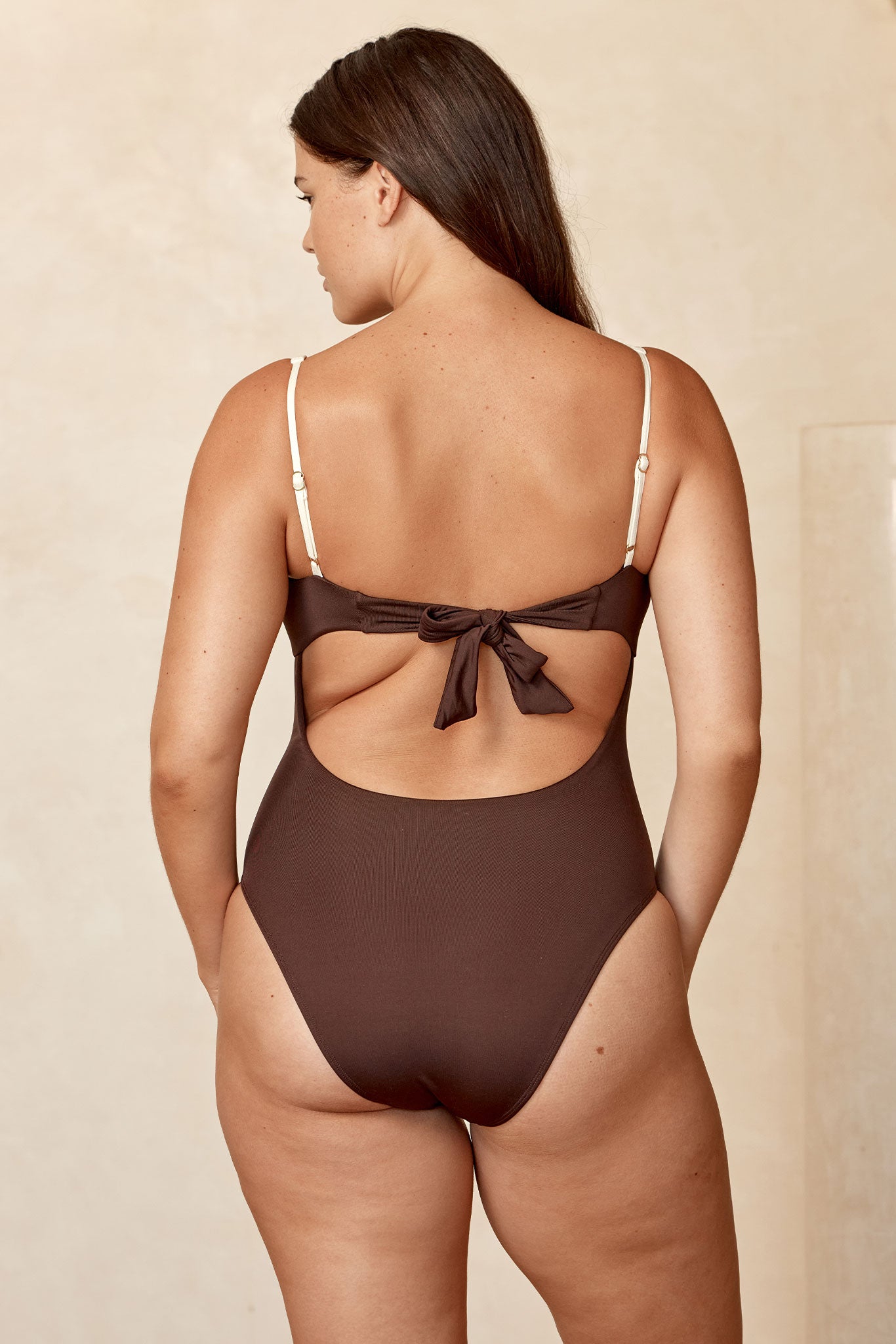 Back view of a woman wearing a brown one-piece swimsuit with adjustable cream straps and a tied bow across the back.