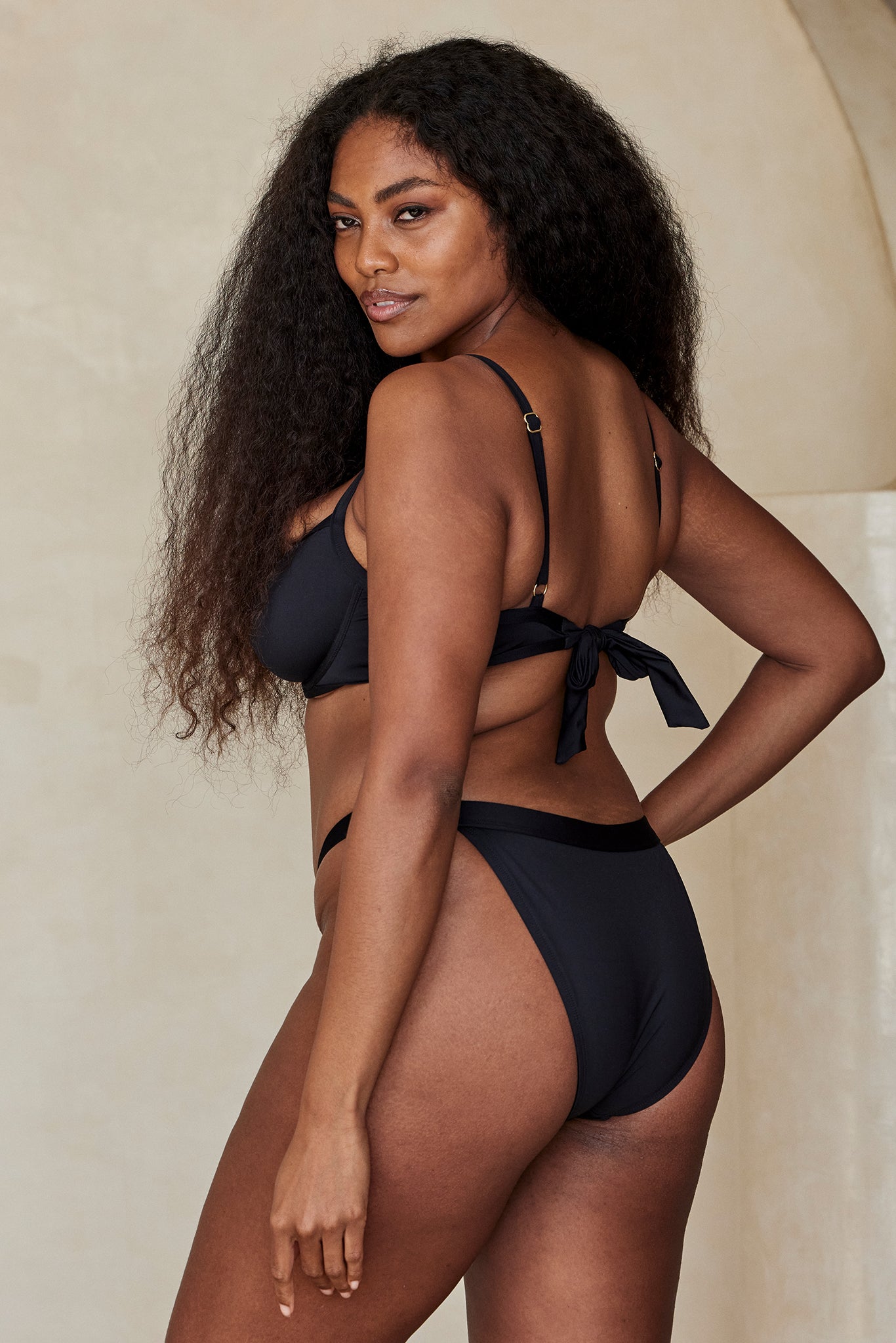 Model wearing black bikini, back three-quarter view with tied bow at back, long curly hair, looking over shoulder.