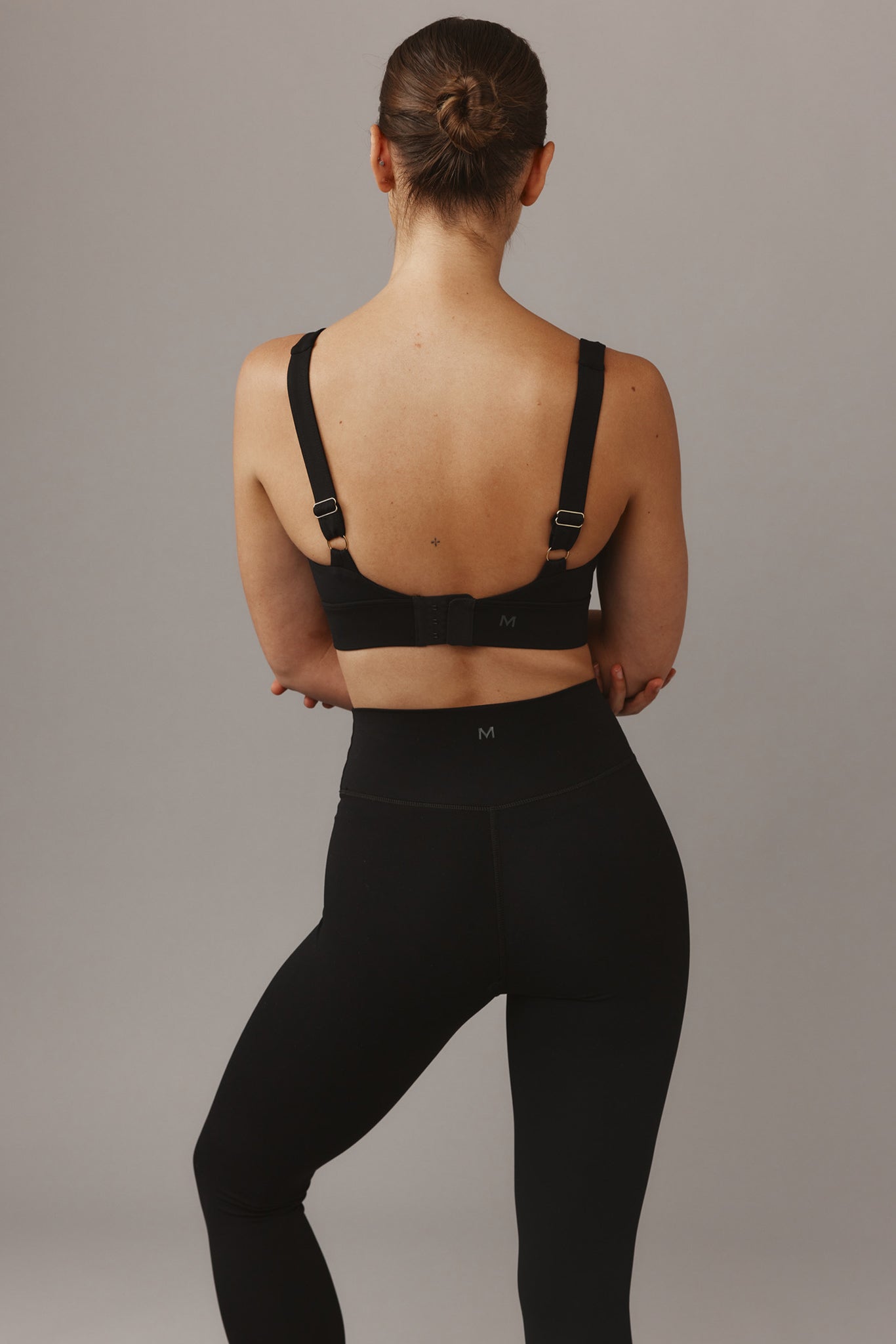 Woman from behind wearing a black sports bra and high-waist leggings with M logos, hair in a bun, small back tattoo