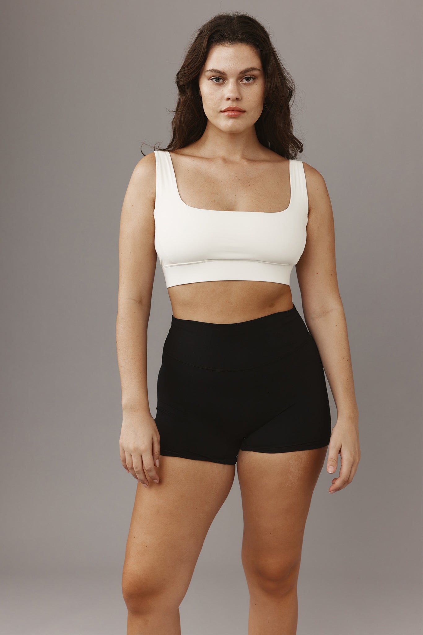 Front-facing woman standing in white square‑neck sports bra and high‑waist black athletic shorts on gray background.
