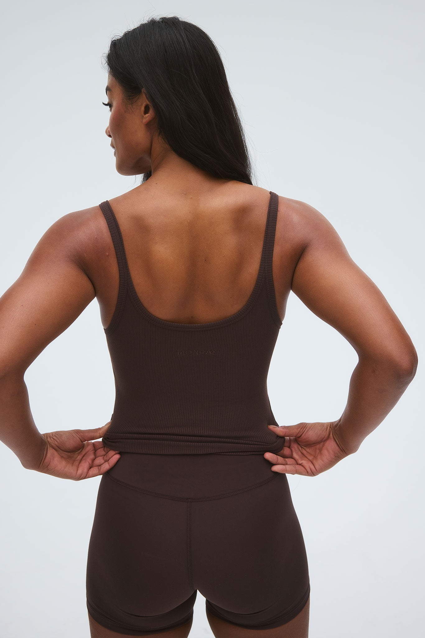 Woman from the back wearing a brown ribbed tank top and matching shorts, pulling down the hem; 'MONDAY' embossed on top.
