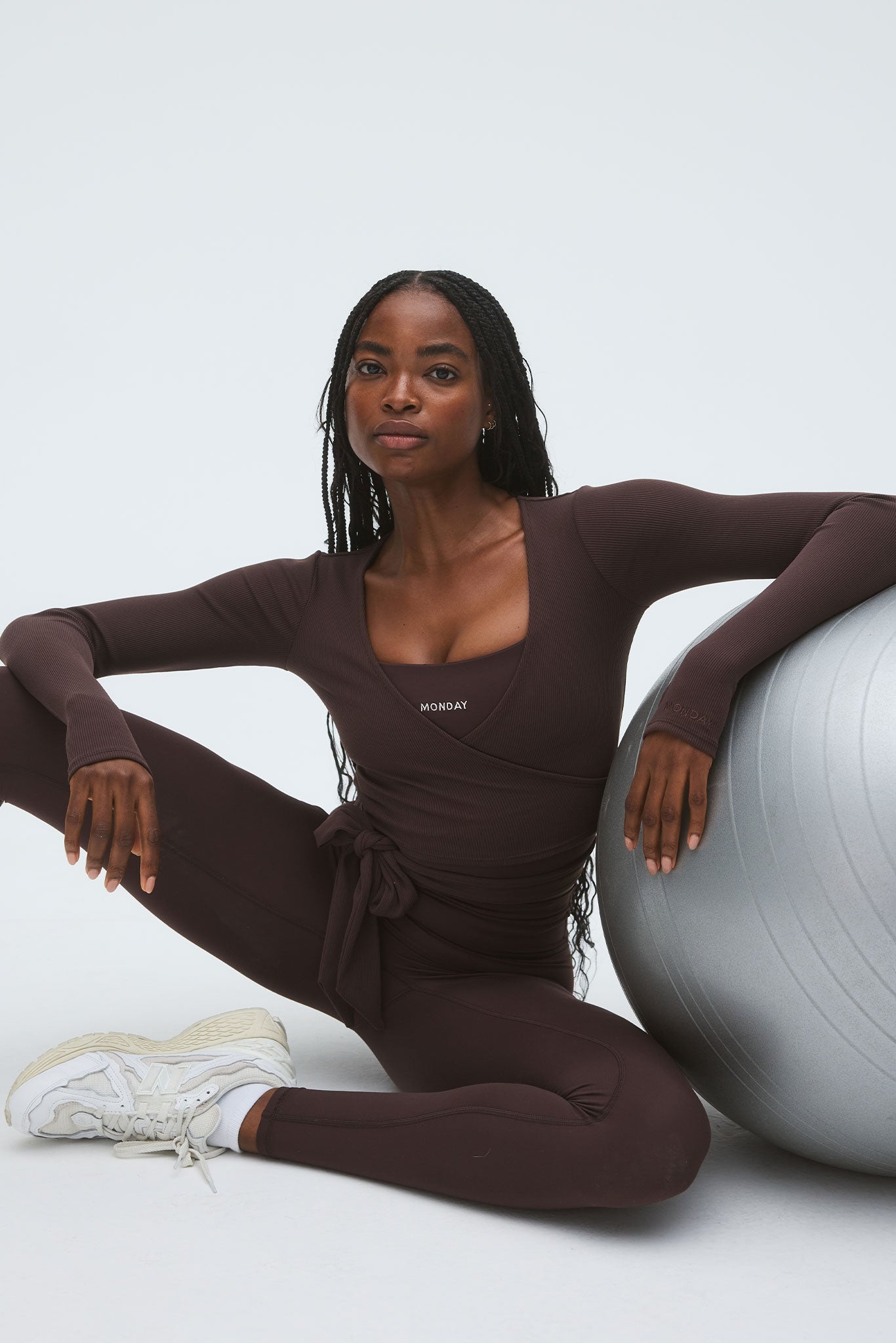 Woman with braided hair in brown wrap workout top and leggings, seated beside a silver exercise ball; 'MONDAY' printed on her top.