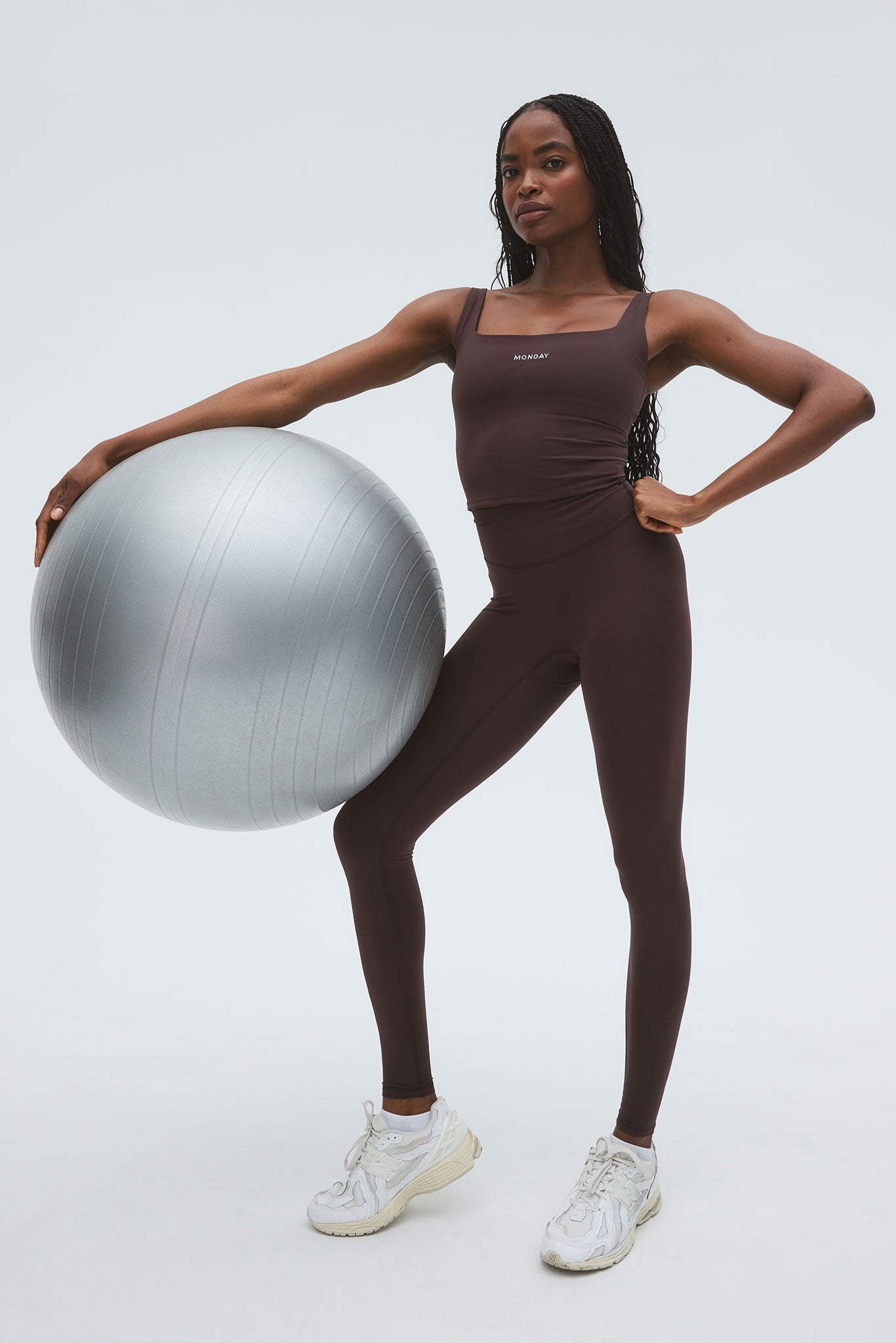 Woman in brown workout set and sneakers holding a silver exercise ball, top labeled 'MONDAY'.