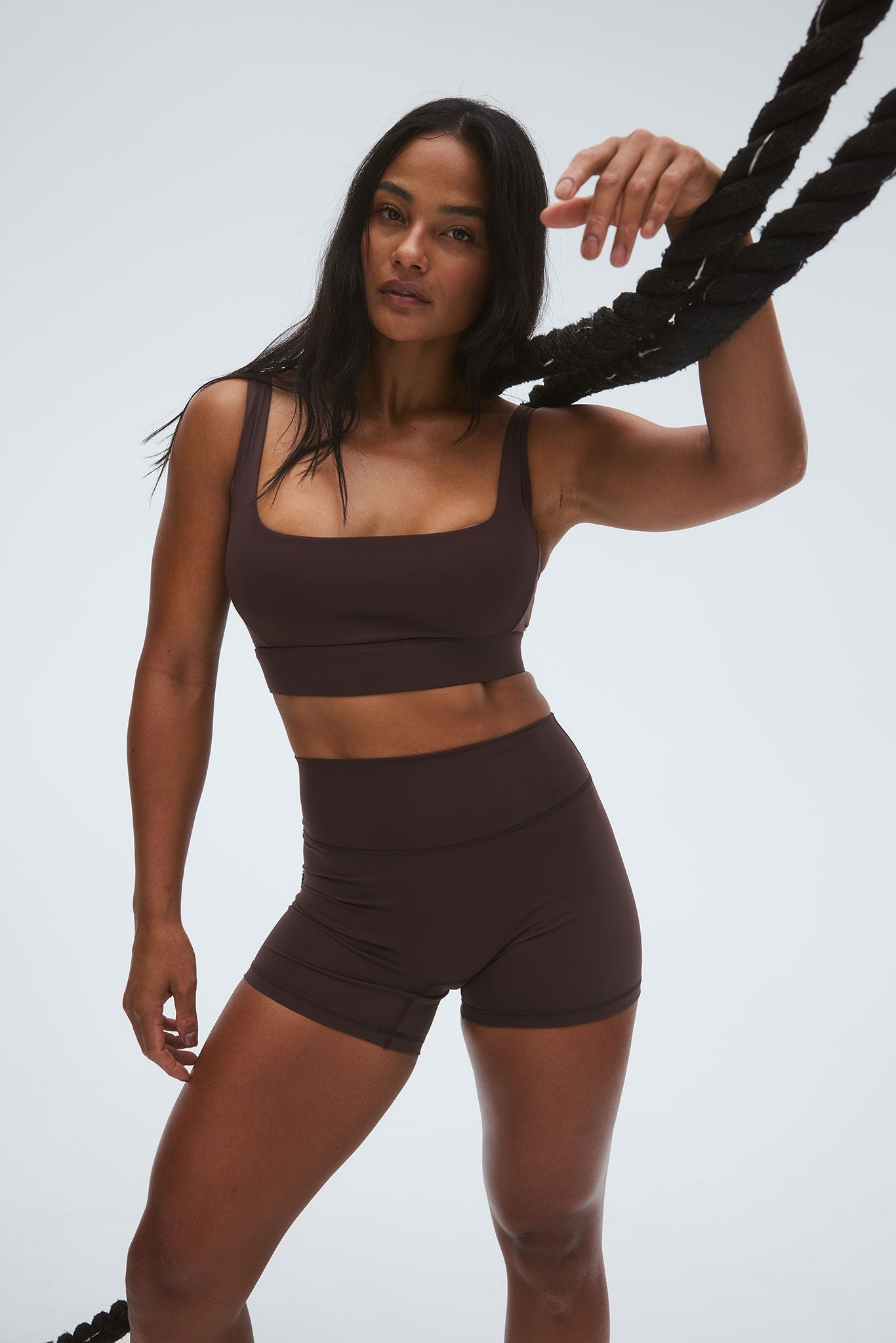 Woman wearing dark brown square-neck sports bra and high-waist fitted shorts, leaning on a thick black rope.