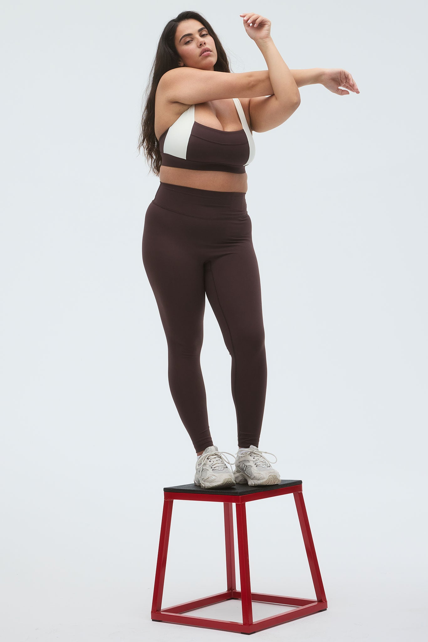 Plus-size woman stretching while standing on a red box, wearing brown leggings, two-tone sports bra, and white sneakers.