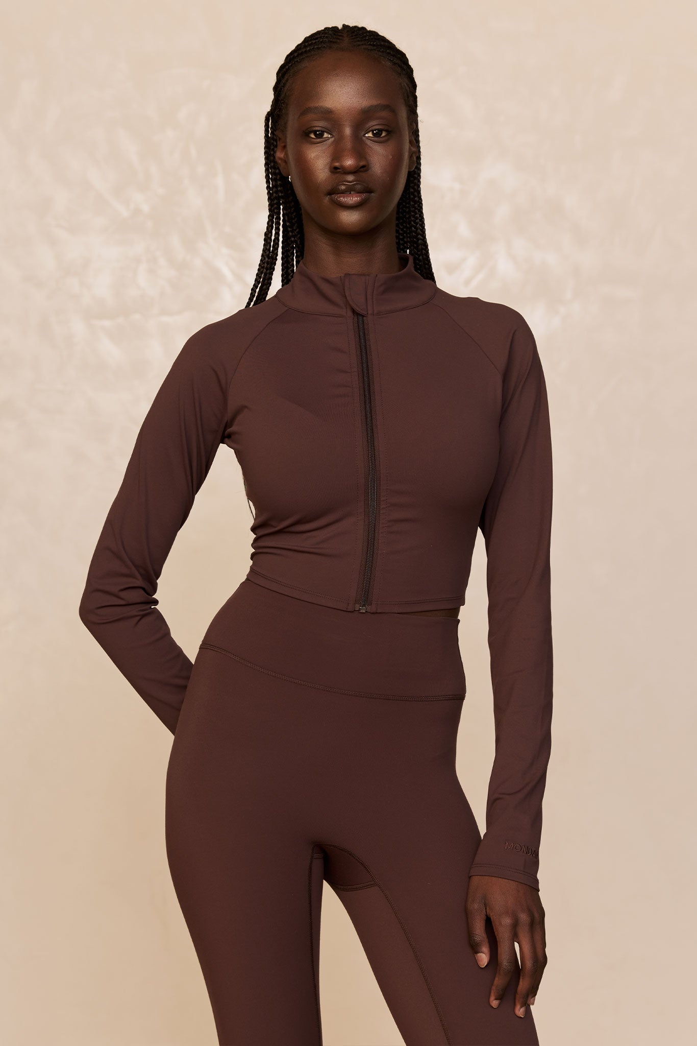 Dark-skinned model wearing chocolate-brown cropped zip jacket and high-waist leggings, MONDAY logo on sleeve, beige backdrop.