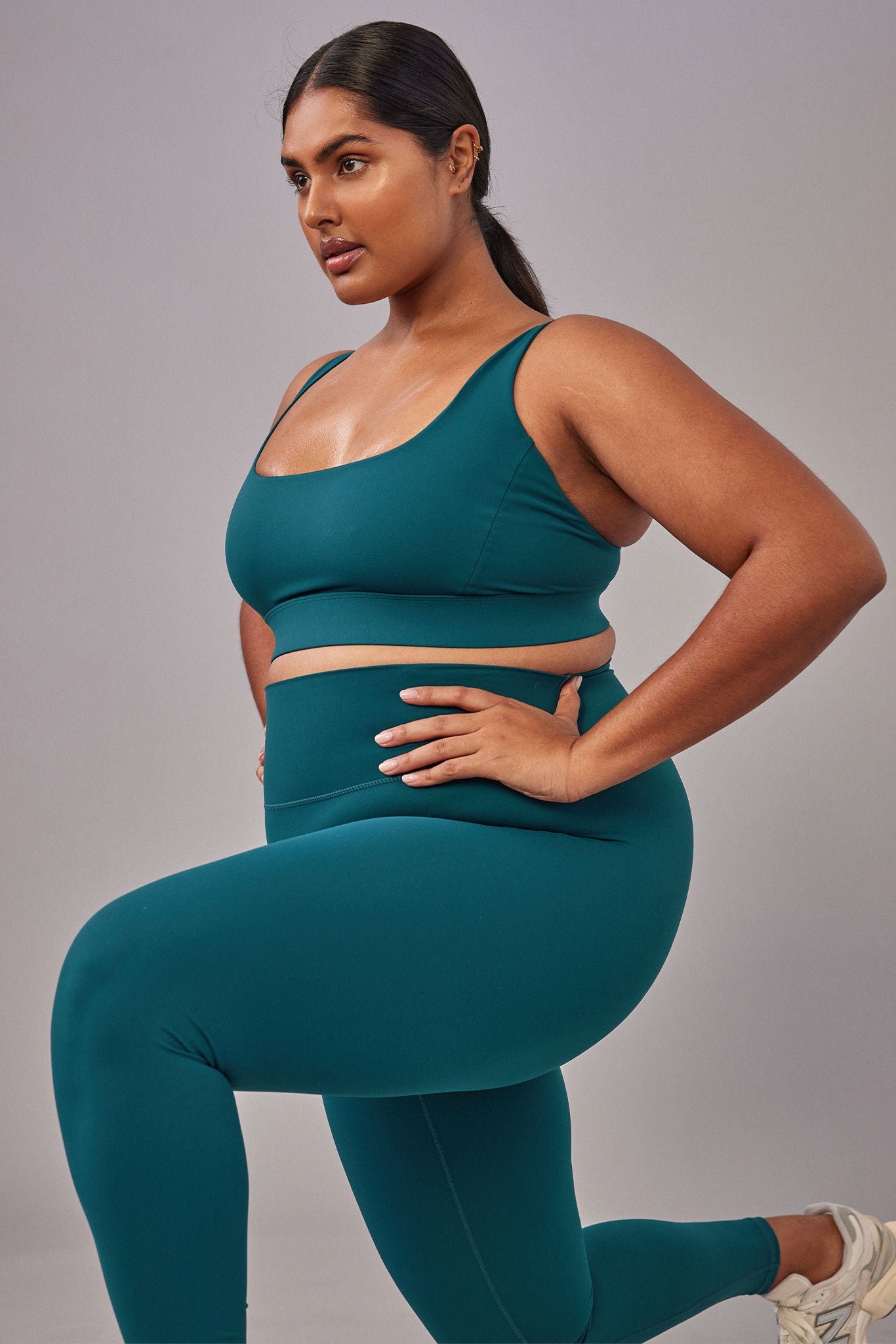 Plus-size woman wearing teal sports bra and high-waist leggings in a lunge, hand on hip, wearing white sneakers.