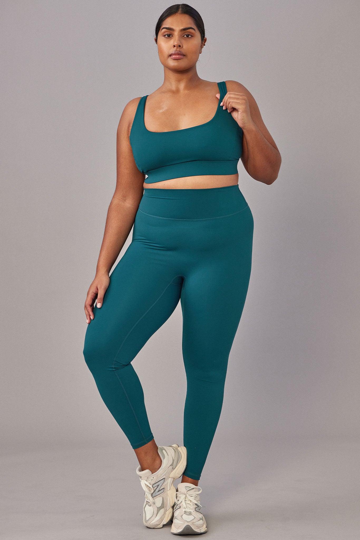 Woman modeling teal sports bra and high-waist leggings with white sneakers, standing with hand on shoulder strap.