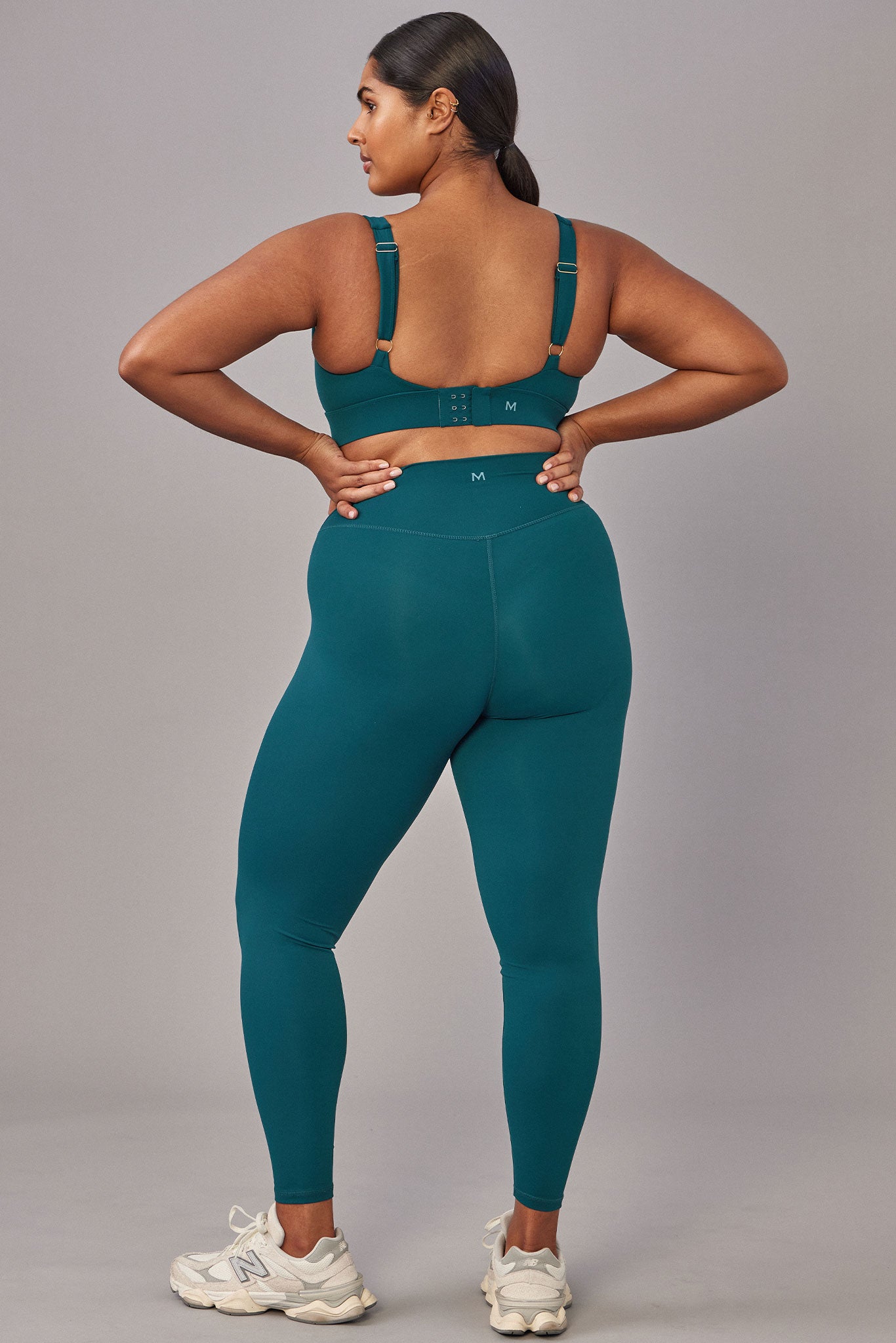 Rear view of person in teal high-waist leggings and matching bra; waistband labeled M; white New Balance sneakers with N