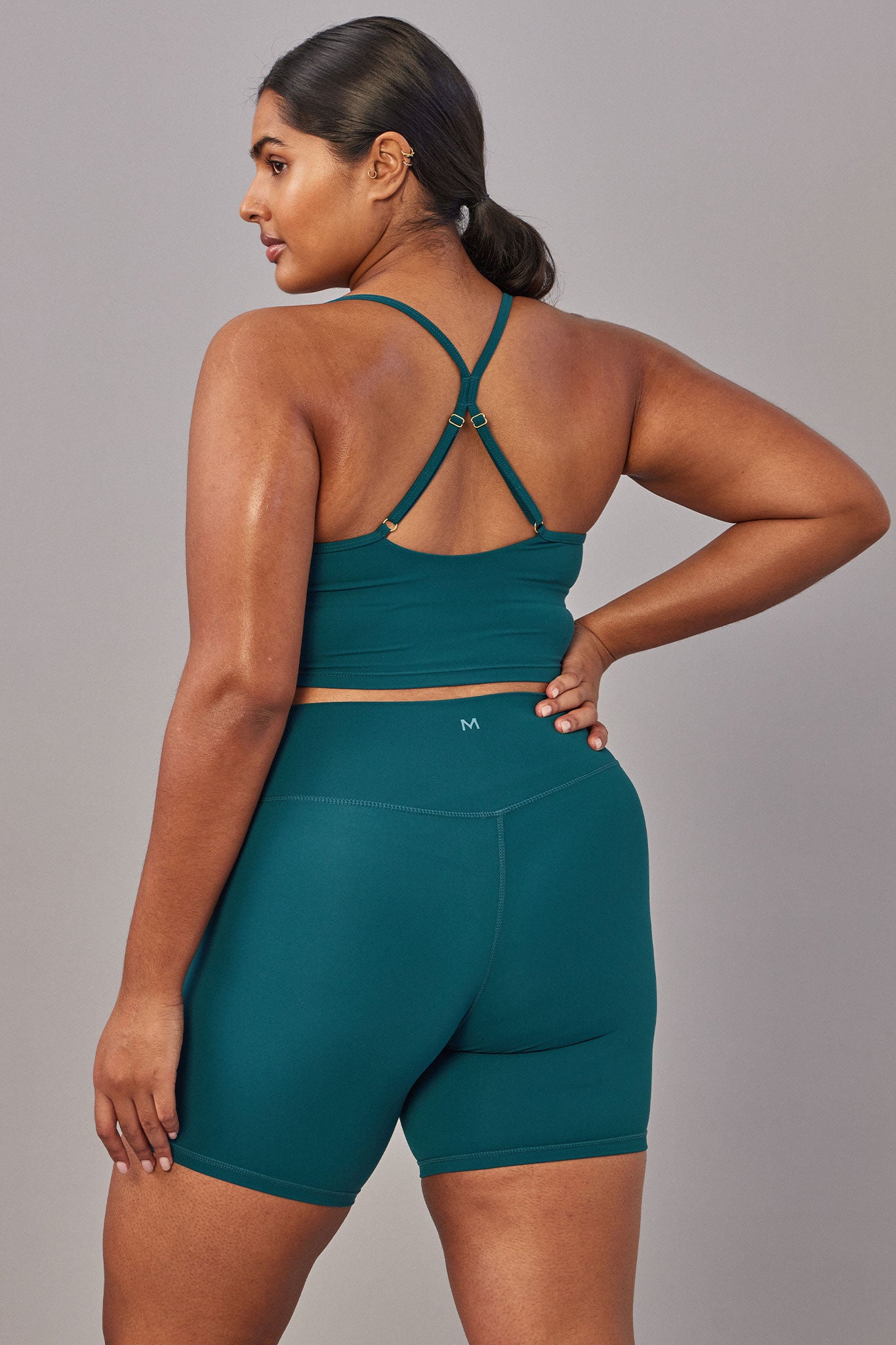Model from back wearing teal racerback sports bra and matching high-waist bike shorts with M logo at center back.