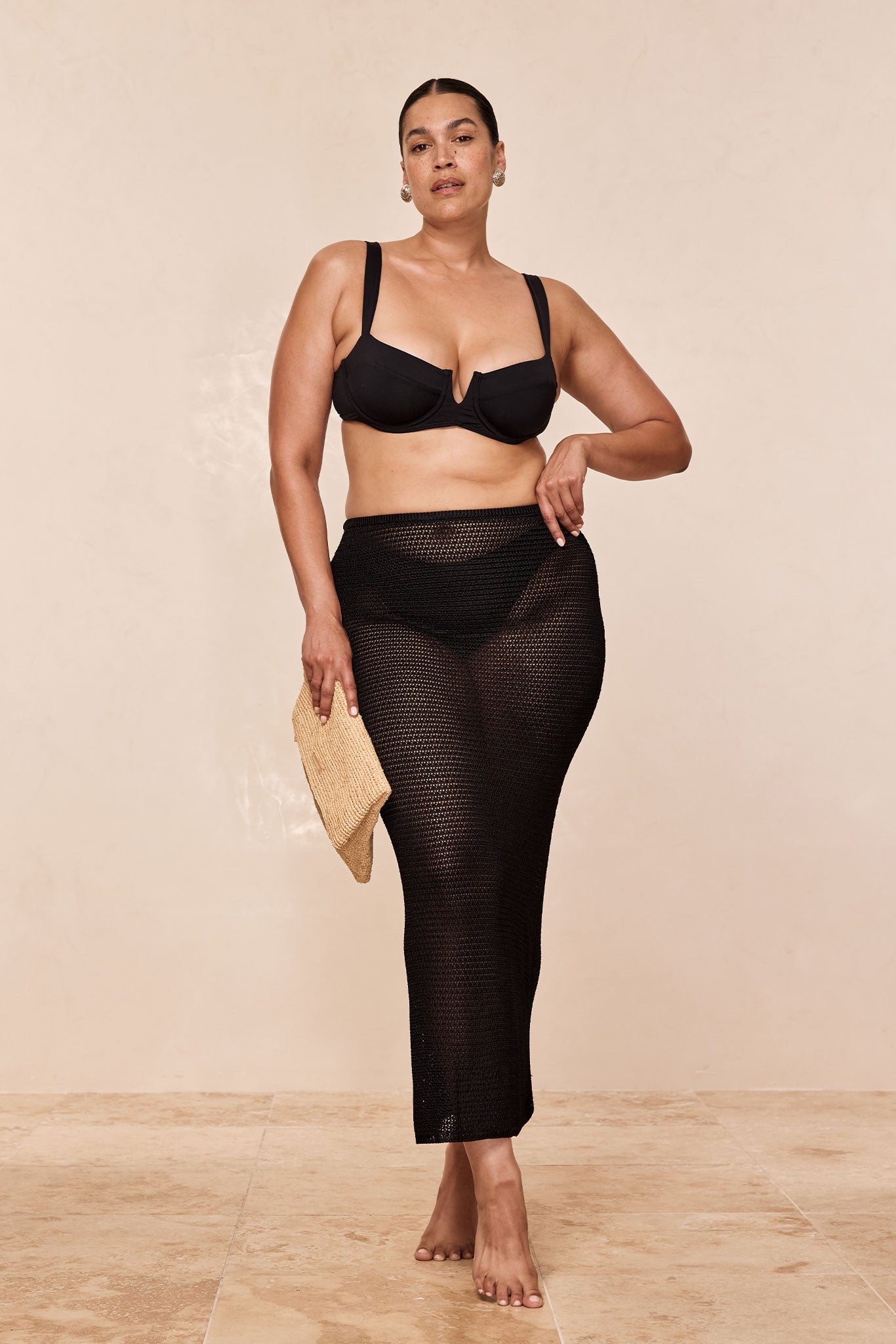 Plus-size model wearing a black underwire bikini top and black sheer knit maxi skirt, holding a woven clutch, barefoot.