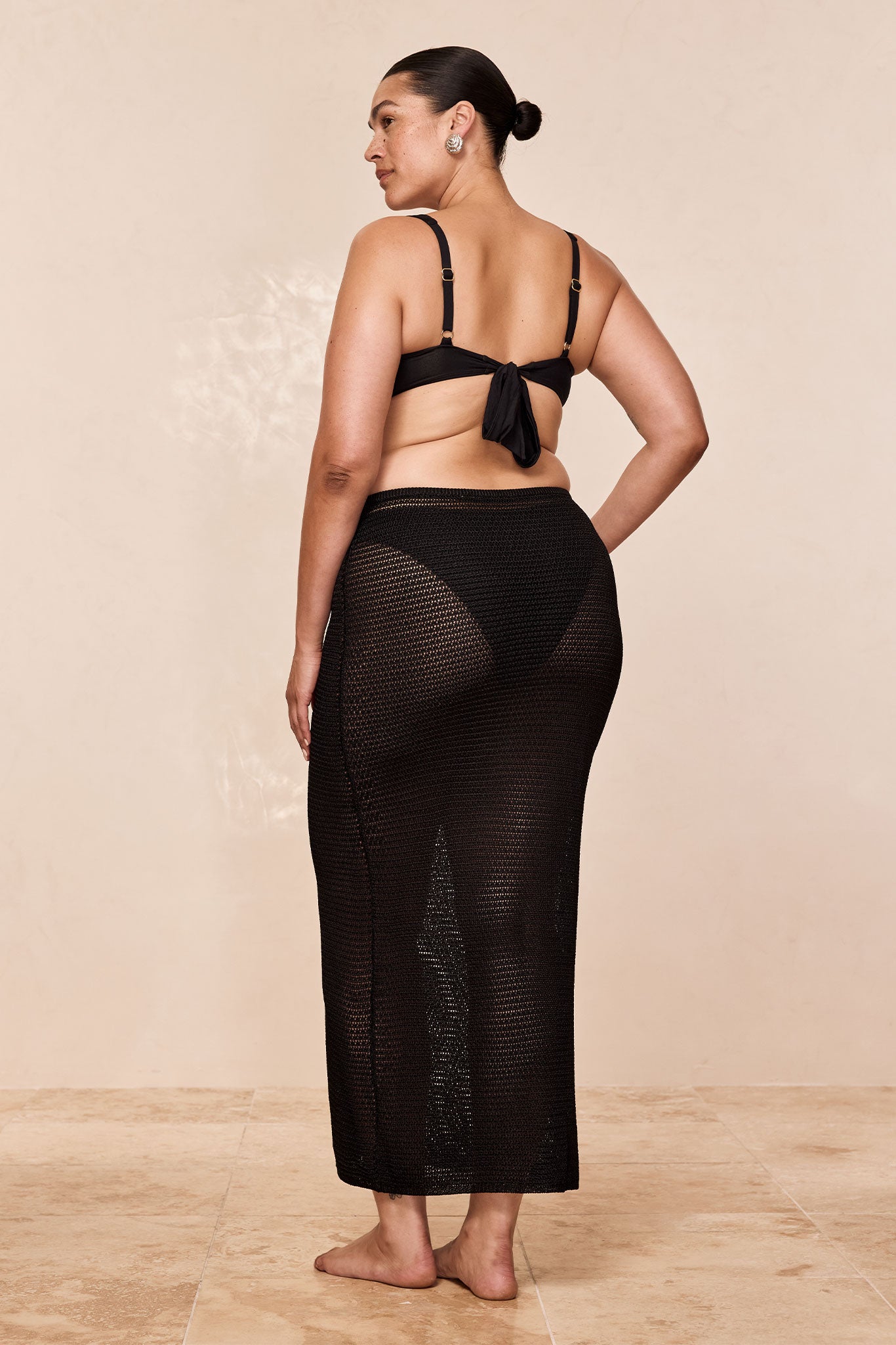 Woman seen from back wearing black bikini top with tied bow and sheer black crochet maxi skirt, barefoot on tiled floor.