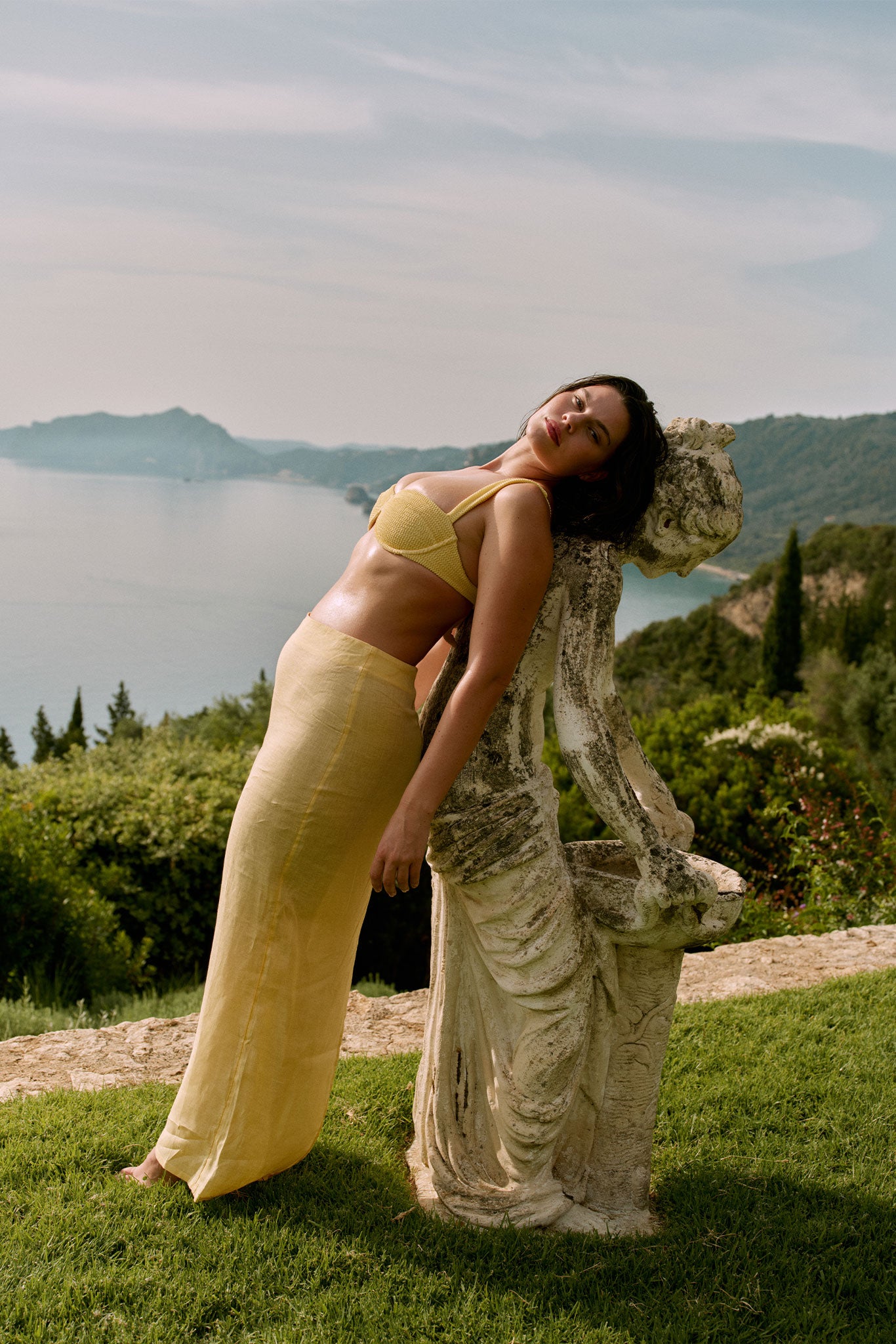 Woman in yellow bikini top and long skirt leaning back against a weathered stone statue on a grassy terrace overlooking the sea.