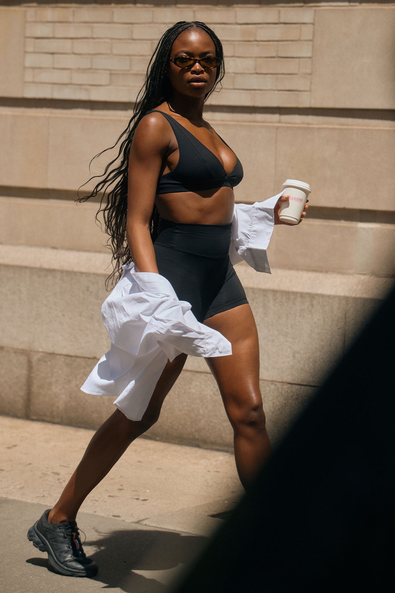 Woman in sunglasses walking in black sports bra and high-waist bike shorts, holding a white coffee cup and a shirt.