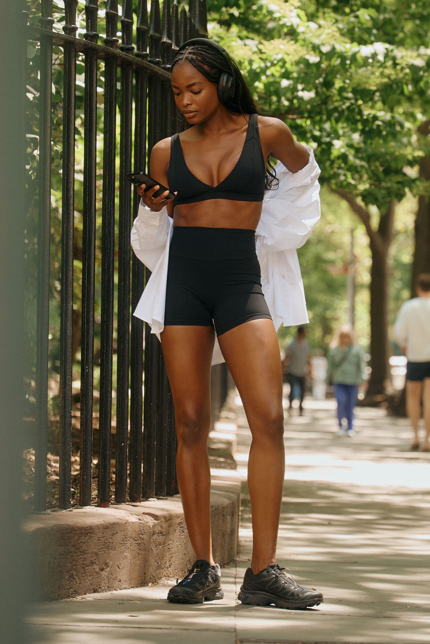 Woman in black sports bra and high-waist shorts with white shirt and headphones, looking at her phone on a shaded sidewalk.