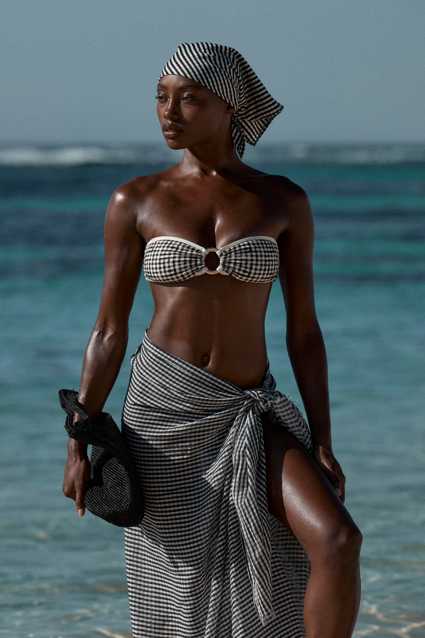 Woman on beach wearing black-and-white gingham bandeau top, matching sarong and headscarf, holding a black woven hat