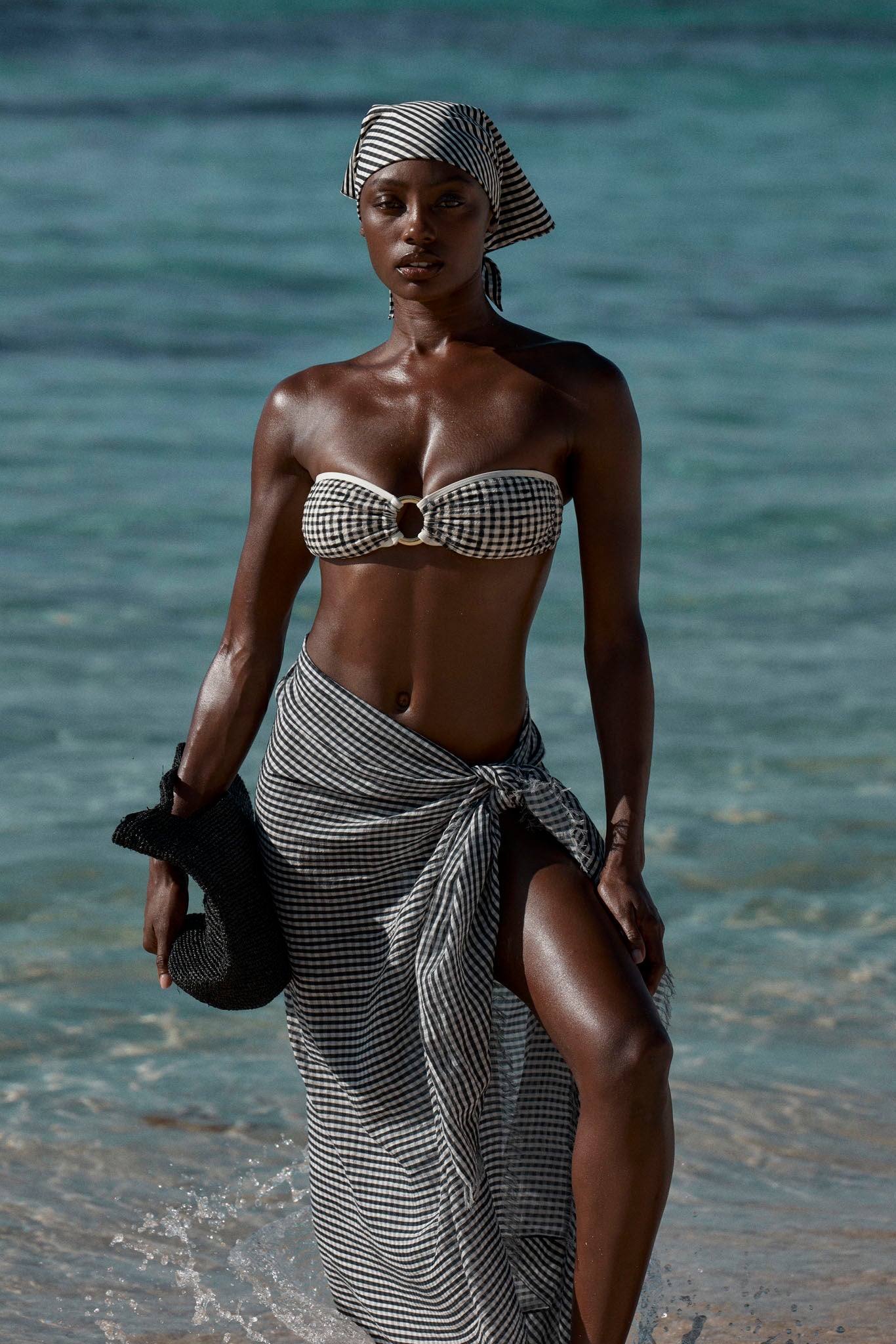 Black woman standing in shallow turquoise sea wearing black-and-white gingham bandeau bikini, matching headscarf and tied sarong, holding a black hat