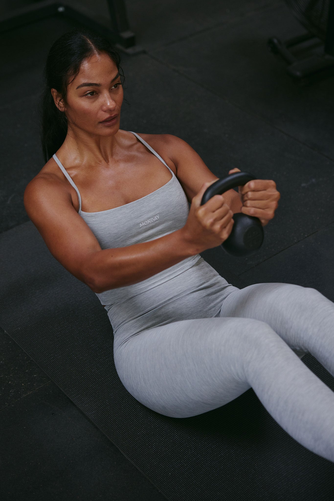 Woman performing a kettlebell Russian twist on a mat, wearing light gray workout set with MONDAY on chest.