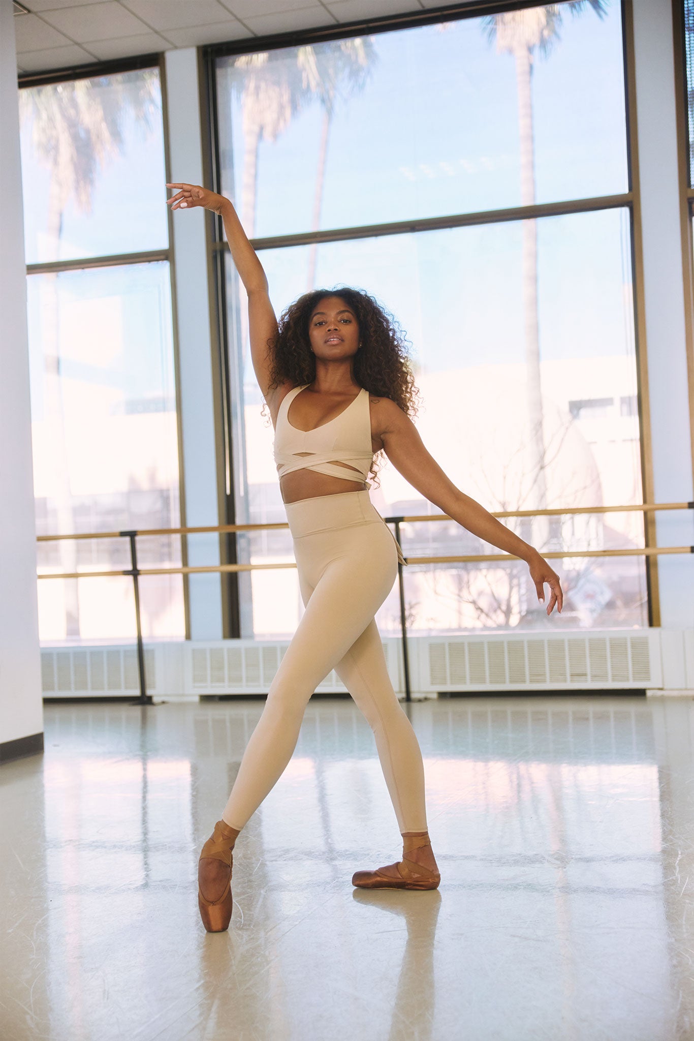 Black ballet dancer en pointe in beige activewear, posing in a sunlit studio with floor-to-ceiling windows and a barre.