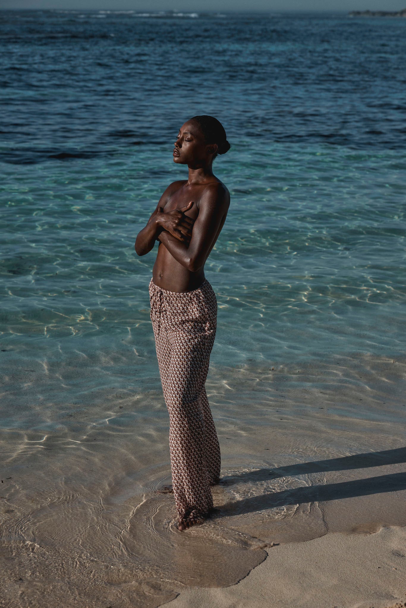 Person standing in shallow sea with patterned pants, arms crossed over chest and eyes closed, sunlight on water.
