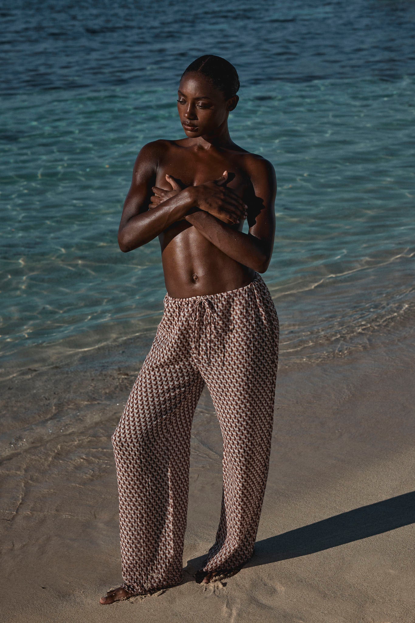 Woman standing barefoot at the shoreline, wearing patterned wide pants and covering her chest with crossed arms.
