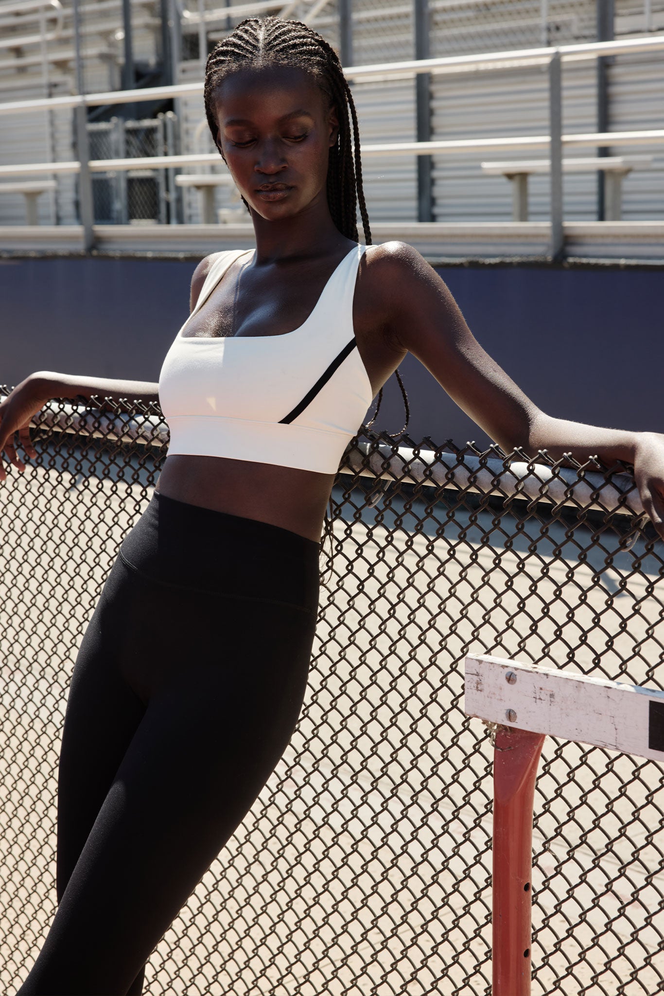 Black woman in white sports bra and black leggings leaning on a chain-link fence at an outdoor track