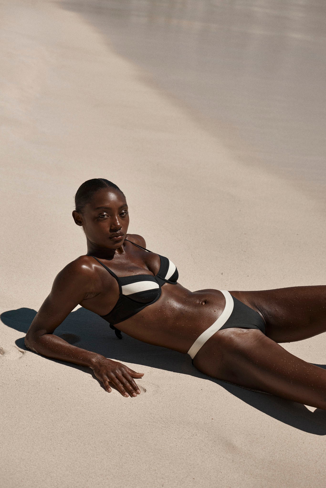 Woman reclining on sunlit sand wearing a black bikini with white trim, relaxed pose, skin glistening.