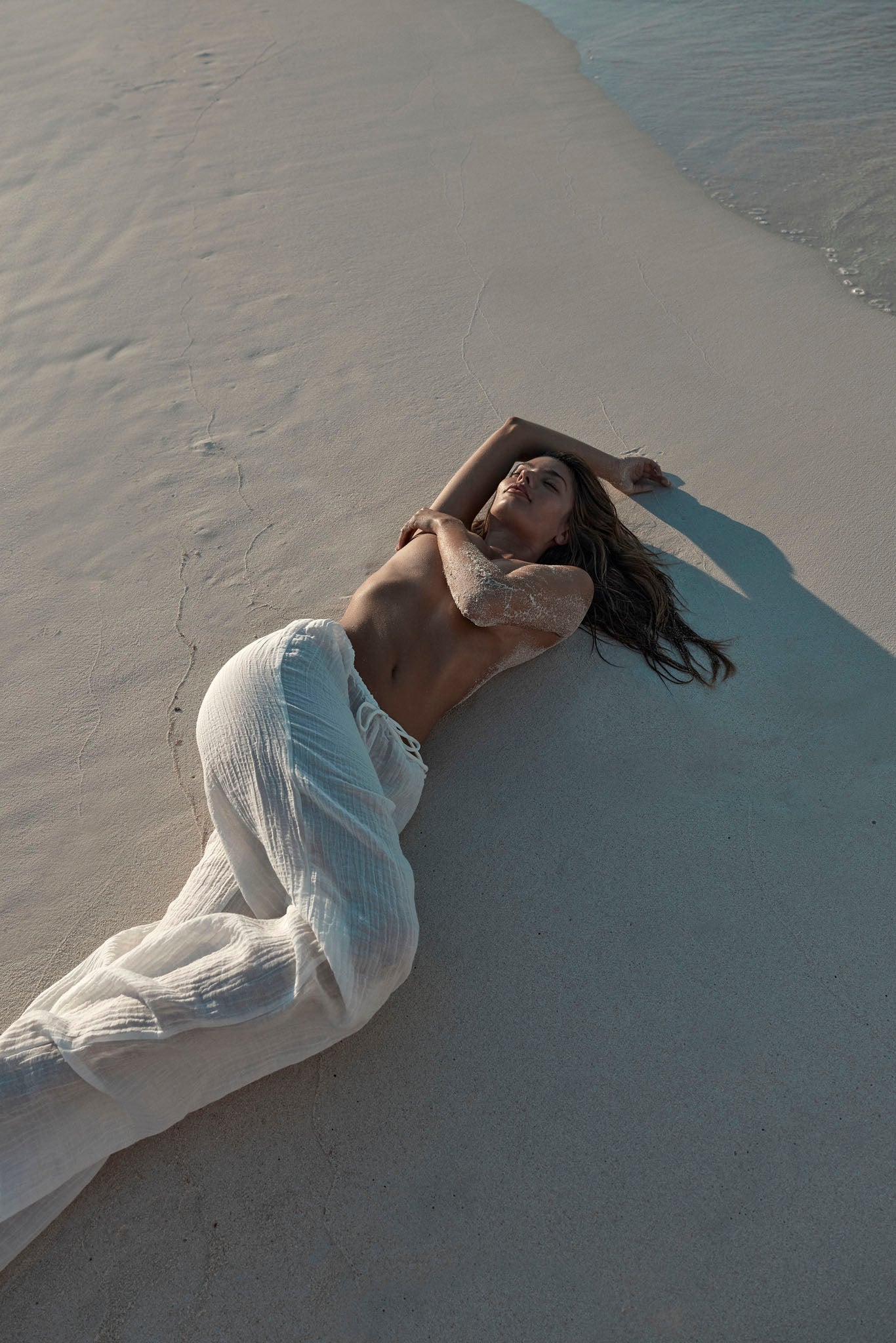 Woman lying topless on white sand near shoreline, eyes closed, wearing sheer white pants with sand on her arm.