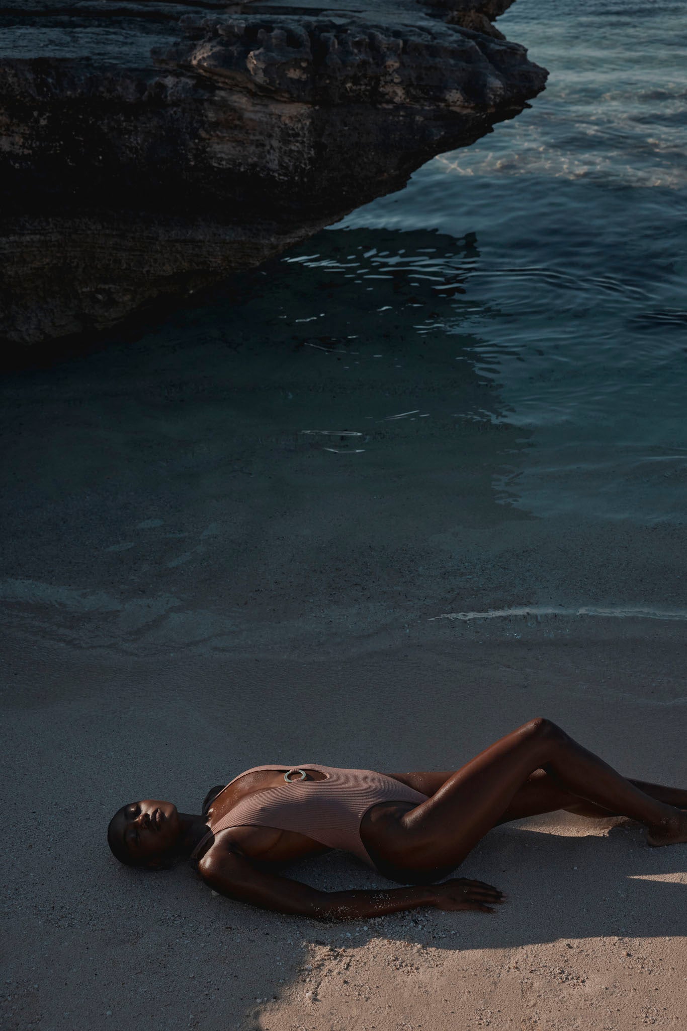 Person in a one-piece swimsuit lying on wet sand beside dark coastal water under an overhanging rock