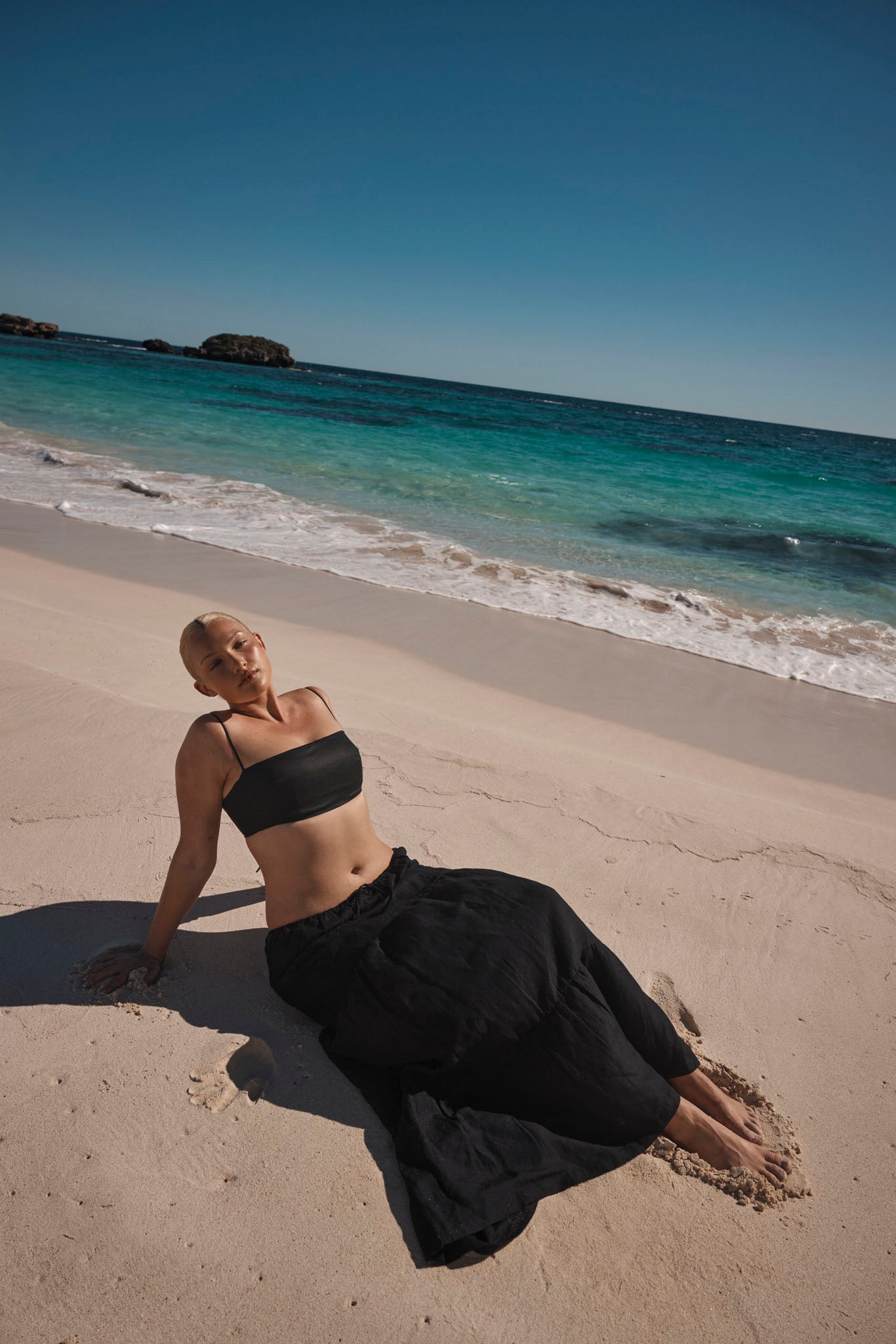 Person reclining on sunlit sandy beach wearing a black tube top and long black skirt, toes in sand with turquoise sea and rocks beyond.