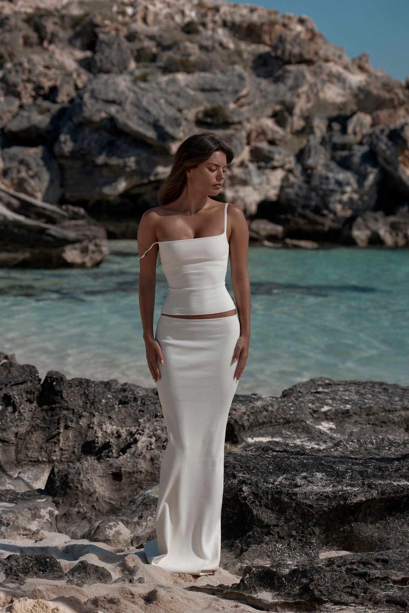 Woman wearing ivory ribbed one-shoulder crop top and matching long ribbed skirt, standing on rocky beach