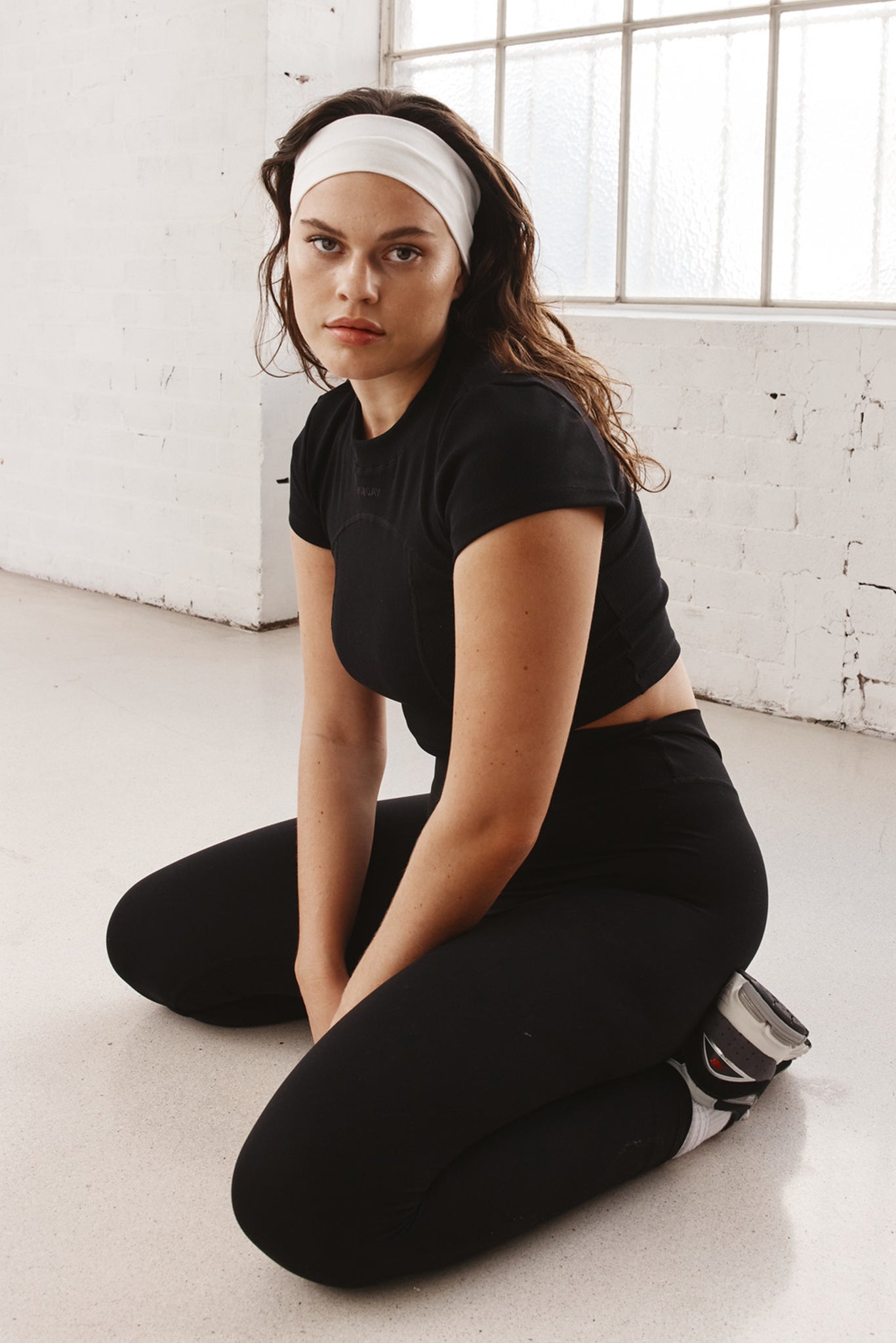 Woman kneeling on studio floor in black workout clothes and white headband, looking toward camera.