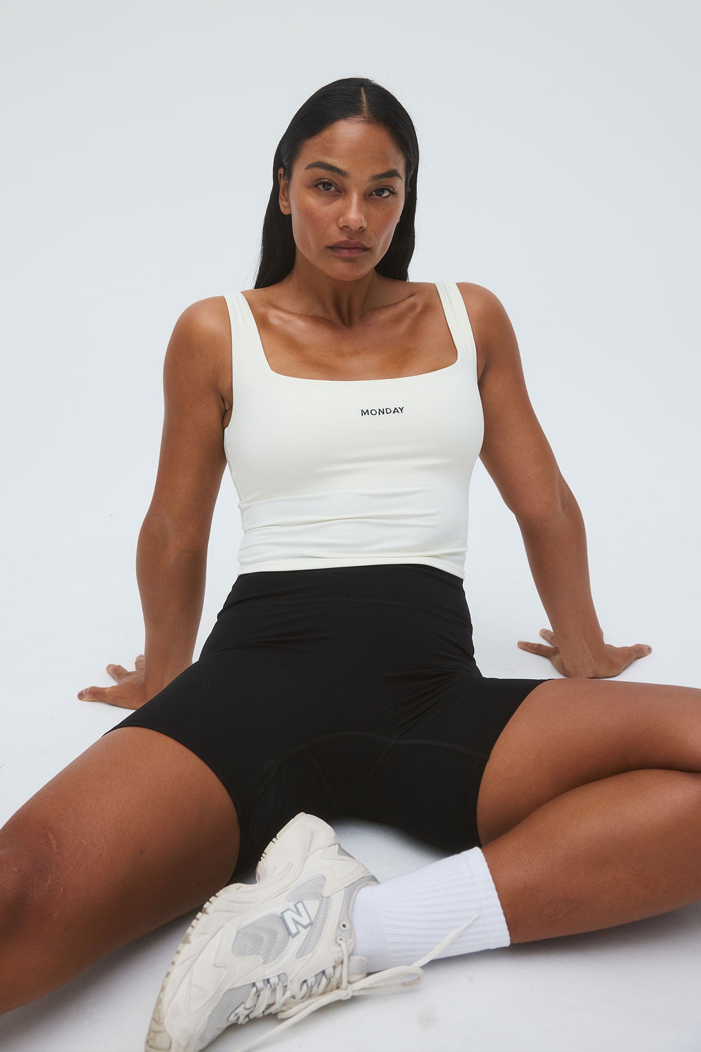 Seated woman wearing white tank top labeled MONDAY, black bike shorts and white New Balance sneakers with 'N' logo