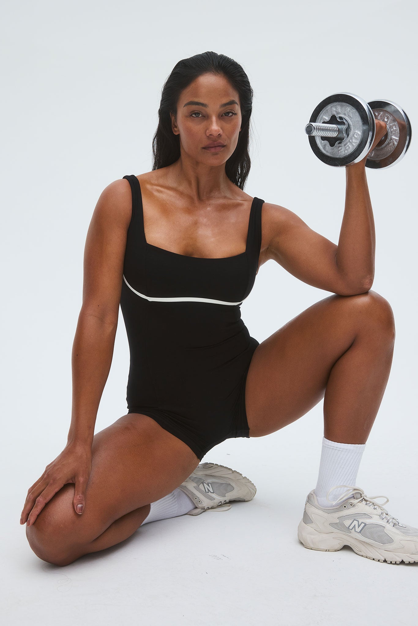 Kneeling woman in black bodysuit holding a dumbbell, wearing white socks and sneakers with N logo