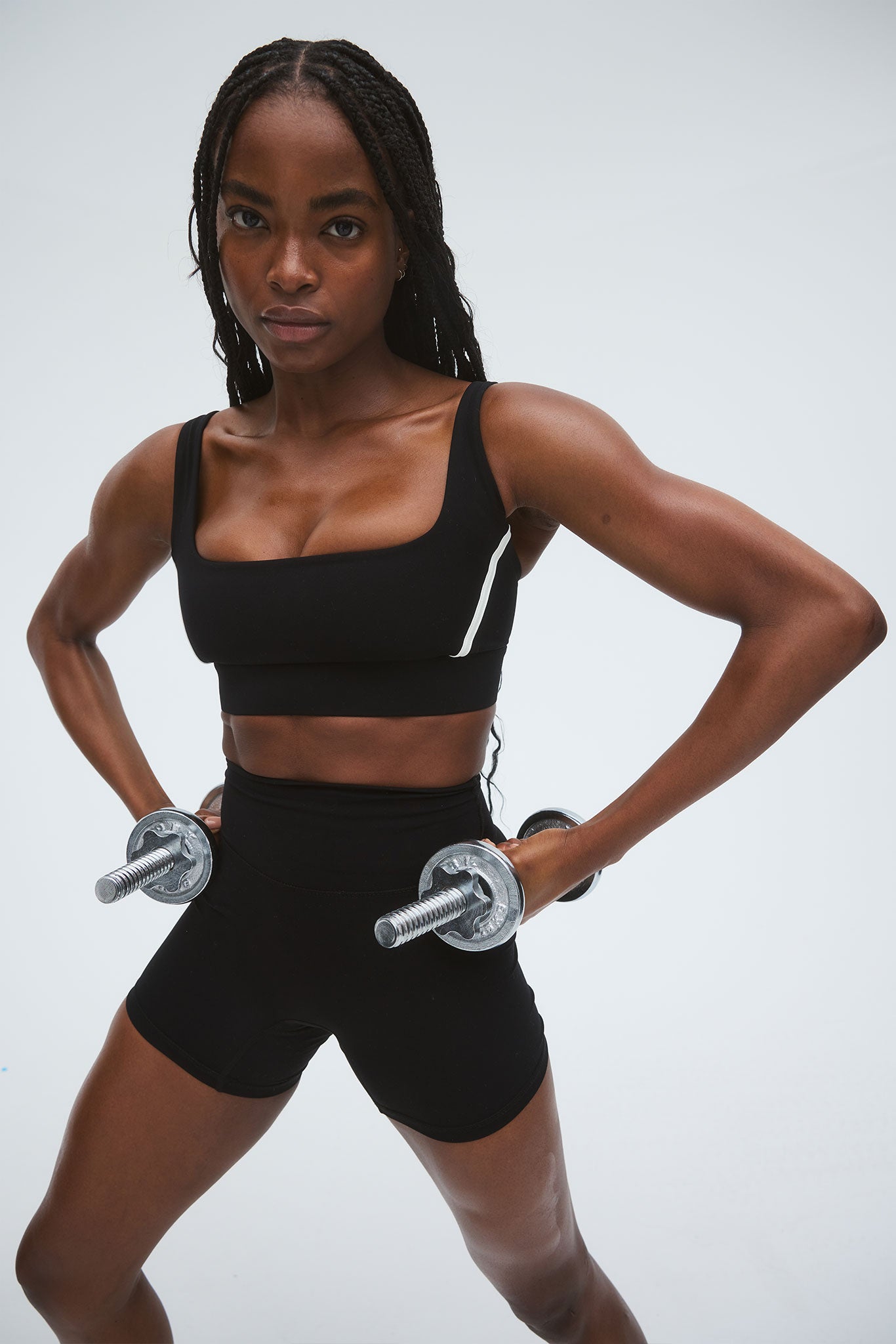 Woman in black sports bra and shorts holding dumbbells at her hips, wide-legged athletic stance, facing the camera