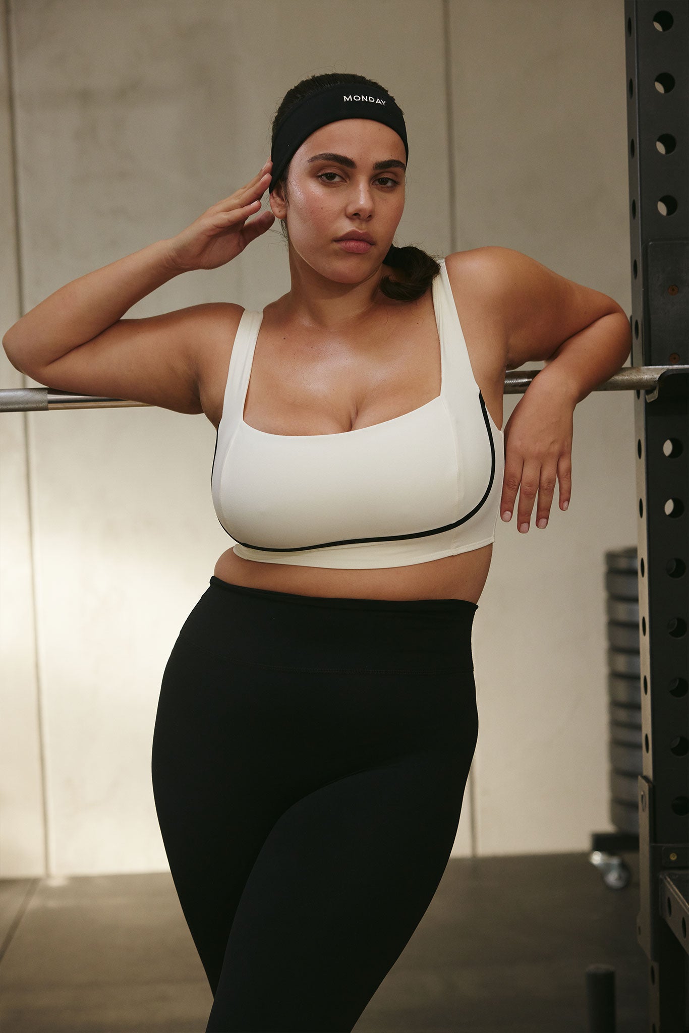 Woman in white sports bra and black leggings leaning on a barbell rack, wearing headband reading MONDAY.
