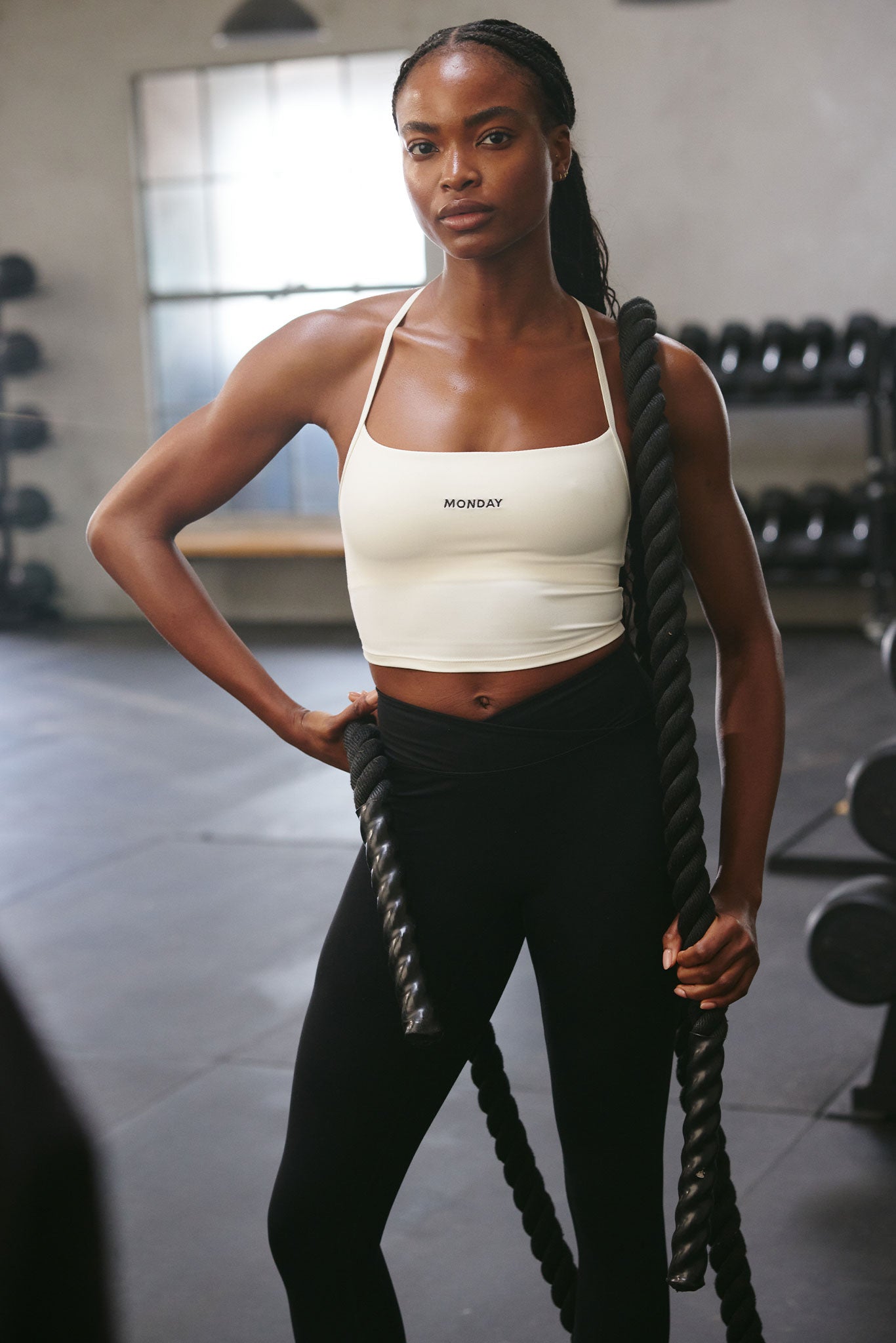 Athletic woman standing in a gym holding battle ropes, wearing a white crop top labeled 'MONDAY'.