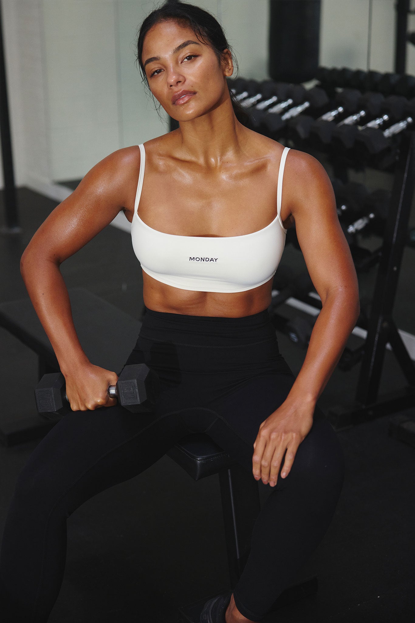 Seated woman in gym wearing white sports bra labeled MONDAY, holding a dumbbell