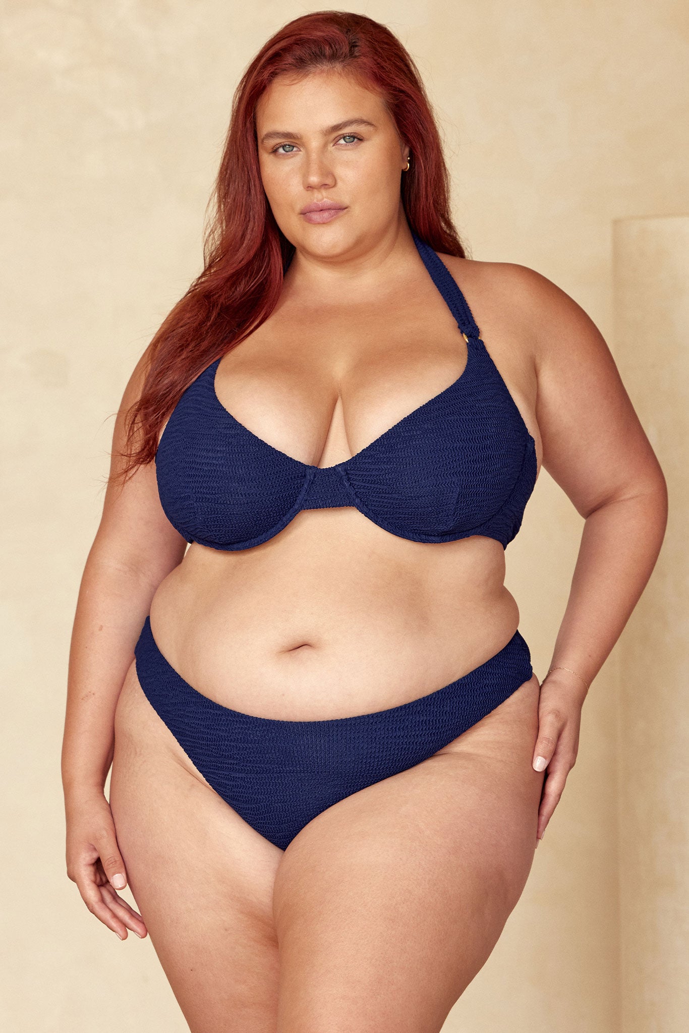 Plus-size woman with red hair wearing a navy textured bikini, front-facing with hands at her sides on a neutral beige background.